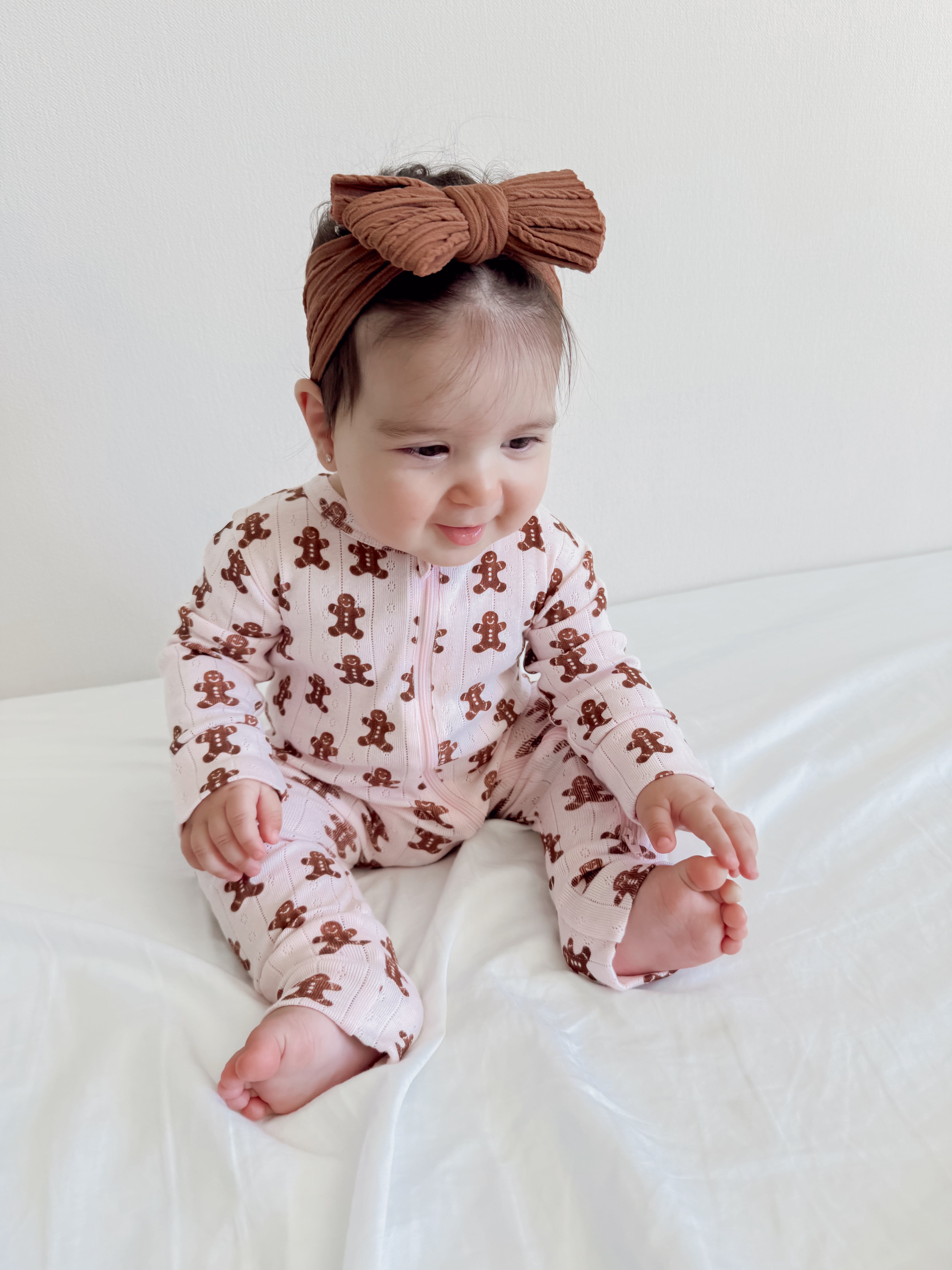 Baby sitting on a white blanket, wearing a gingerbread-patterned outfit and a brown bow headband.