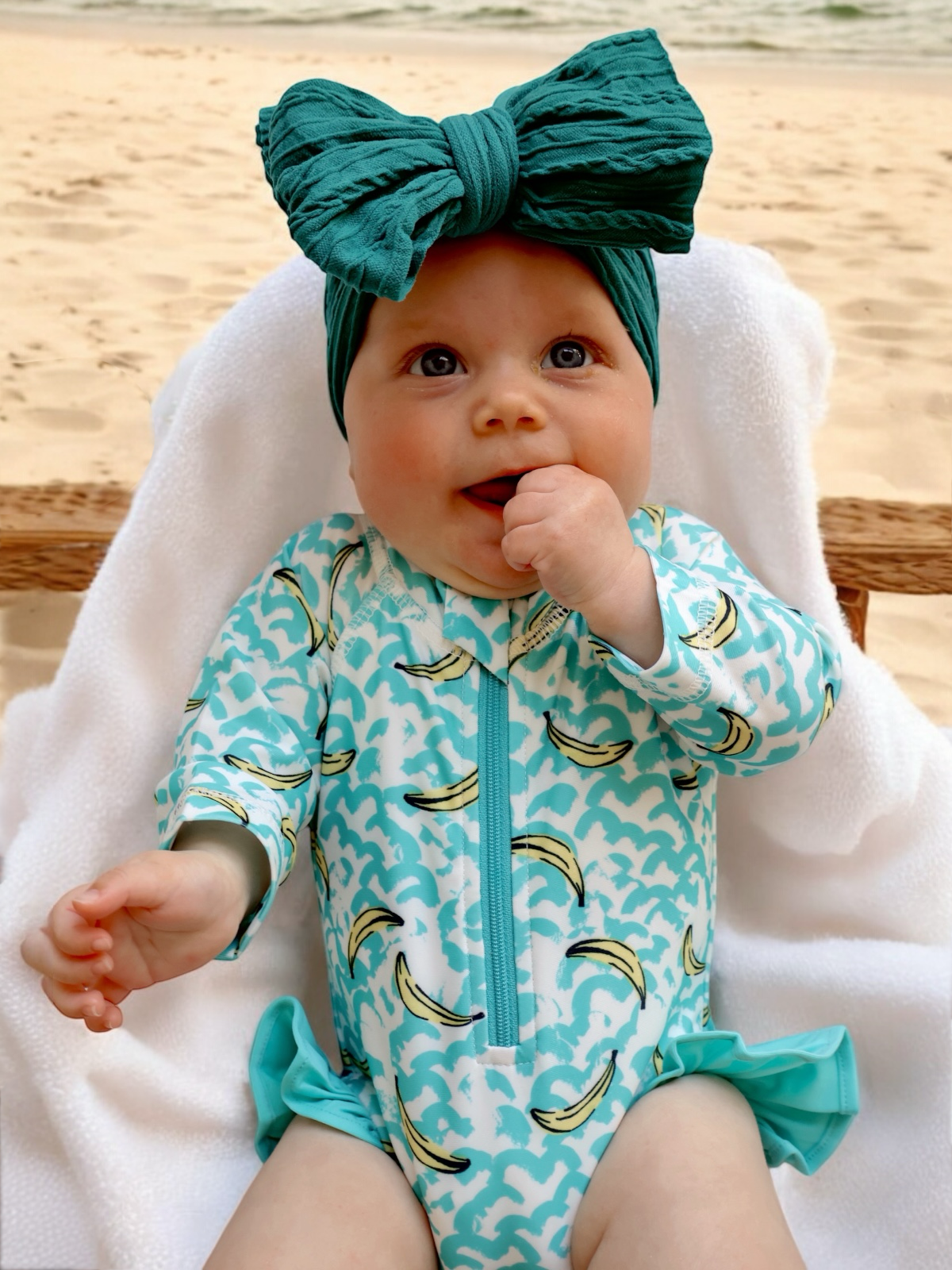Baby in a banana-patterned swimsuit and large teal bow, sitting on a towel by the beach.