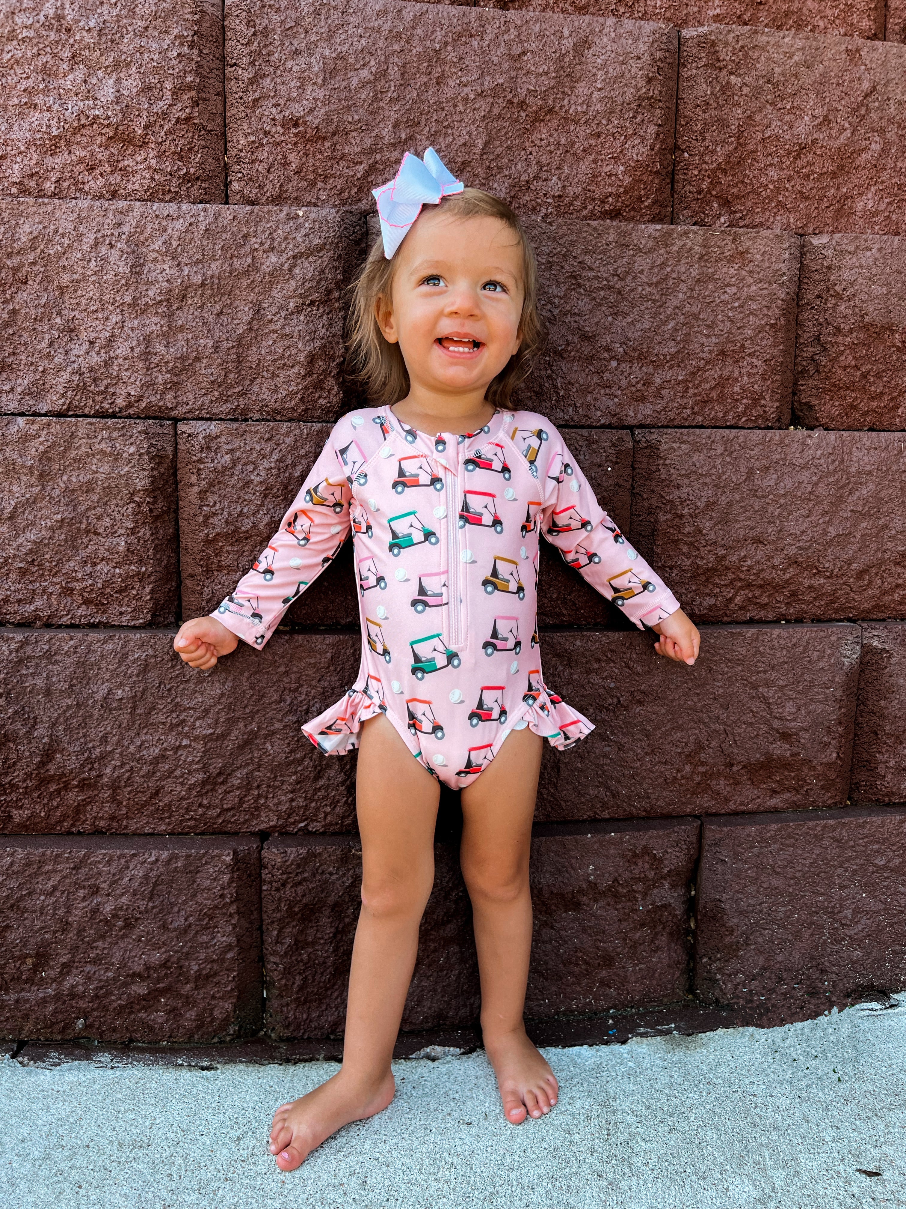 Smiling toddler in a pink swimsuit with a golf cart pattern, standing against a brown stone wall.
