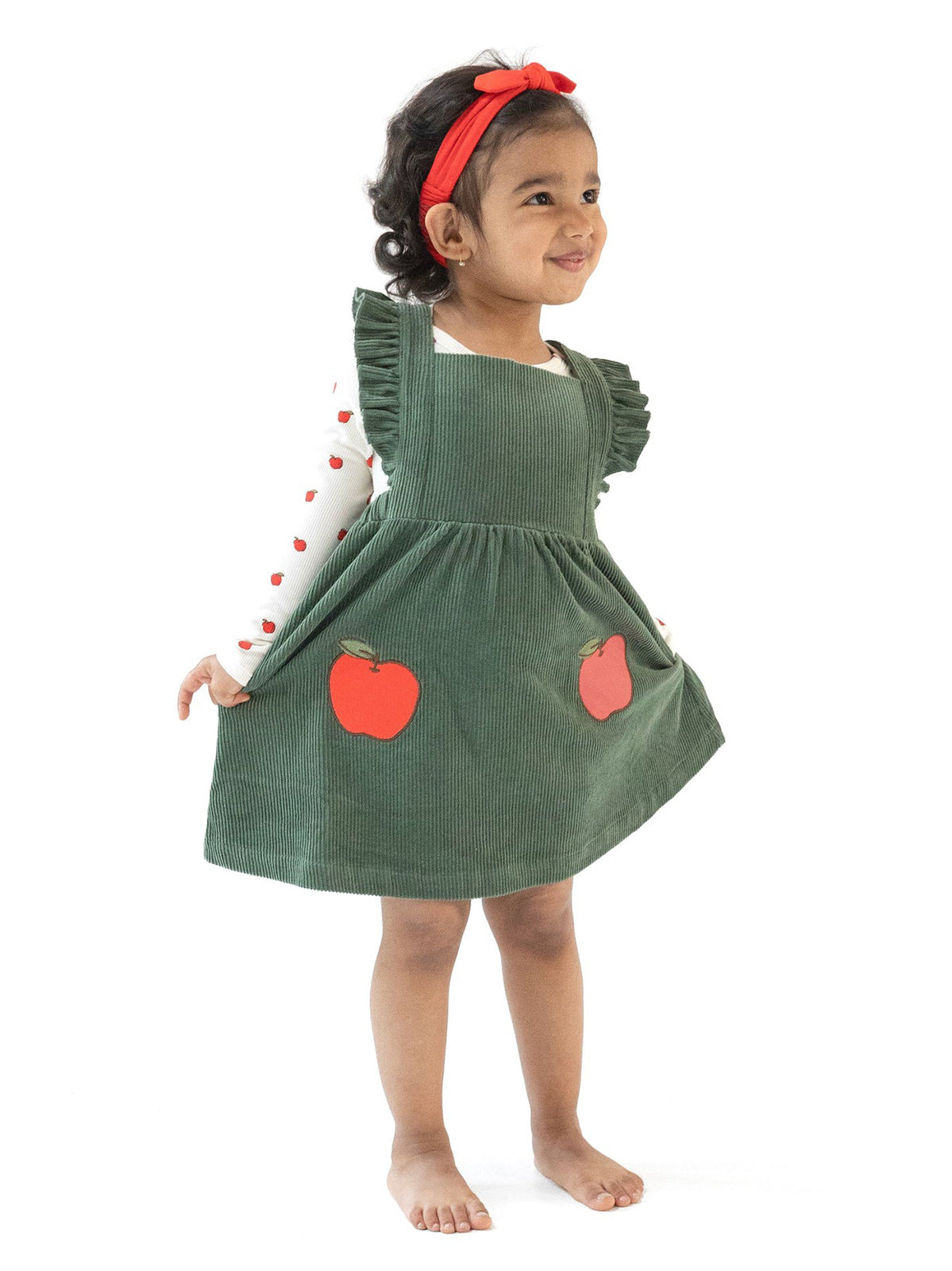 Smiling toddler in a green corduroy dress with apple patches, wearing a red headband and long-sleeve top with hearts.