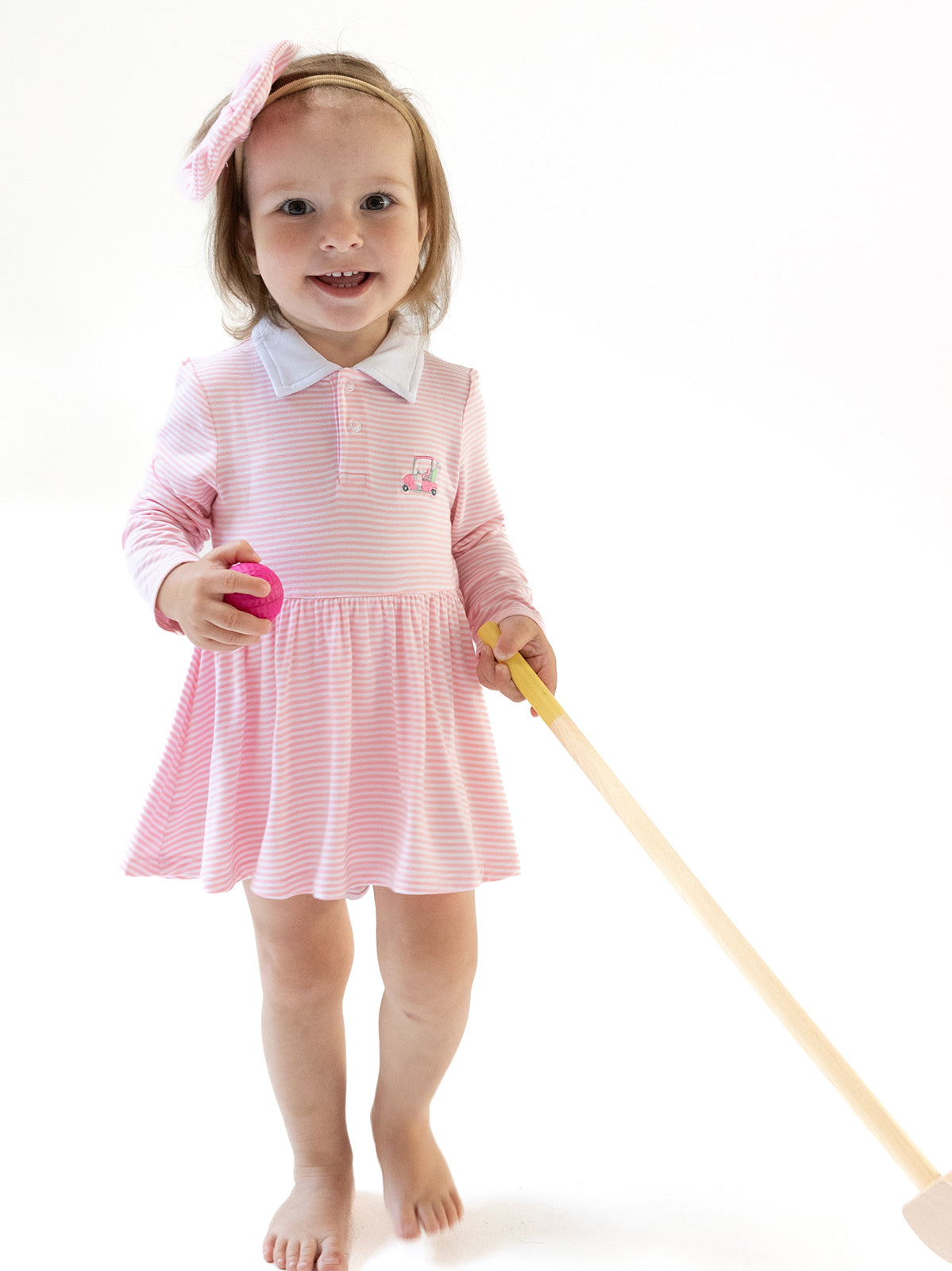 Smiling child in a pink striped dress holding a toy and a wooden stick, standing on a plain white background.