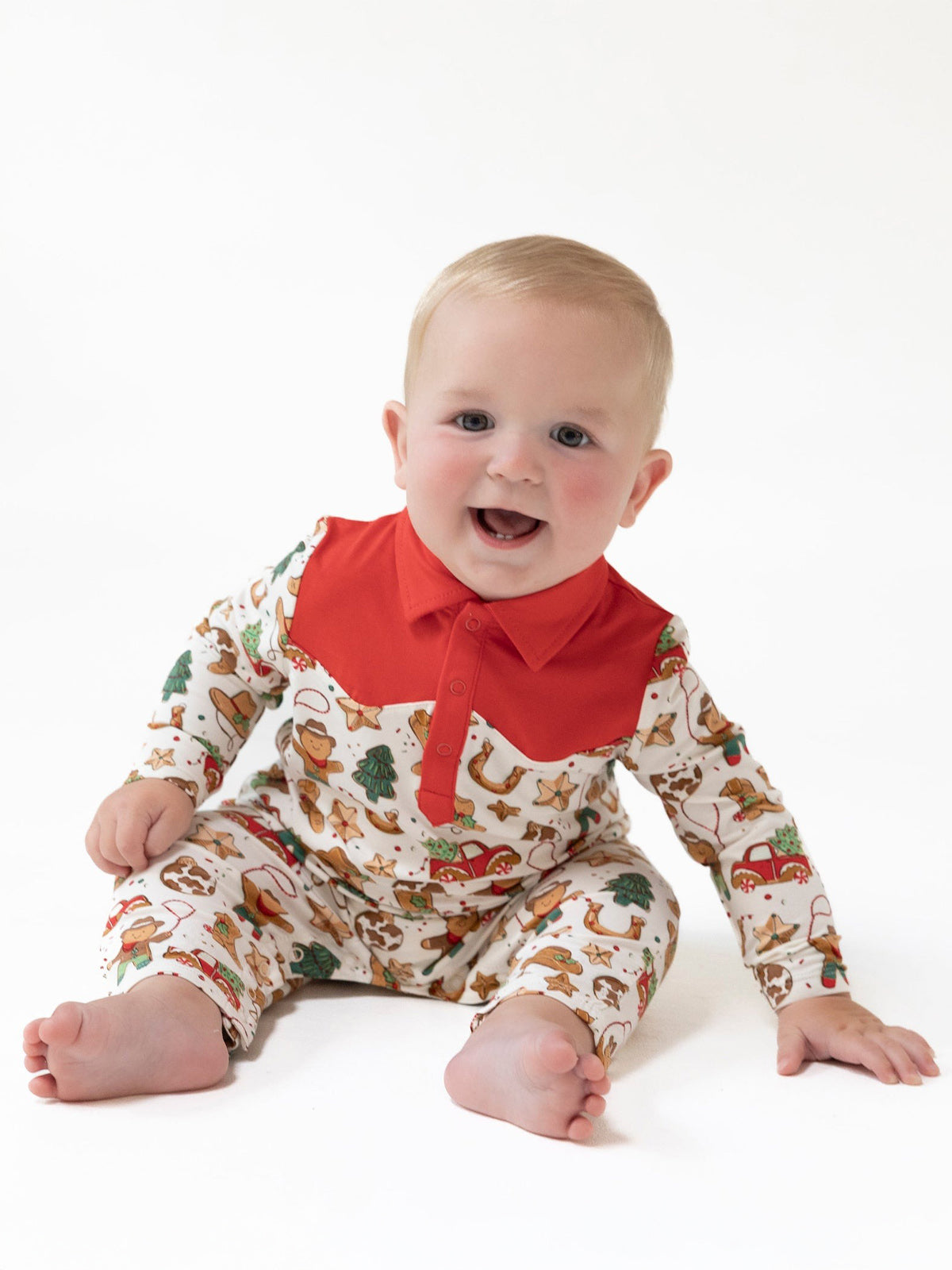 Smiling baby in a festive patterned onesie with a red collar, sitting on a white background.