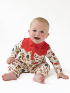 Smiling baby in a festive patterned onesie with a red collar, sitting on a white background.