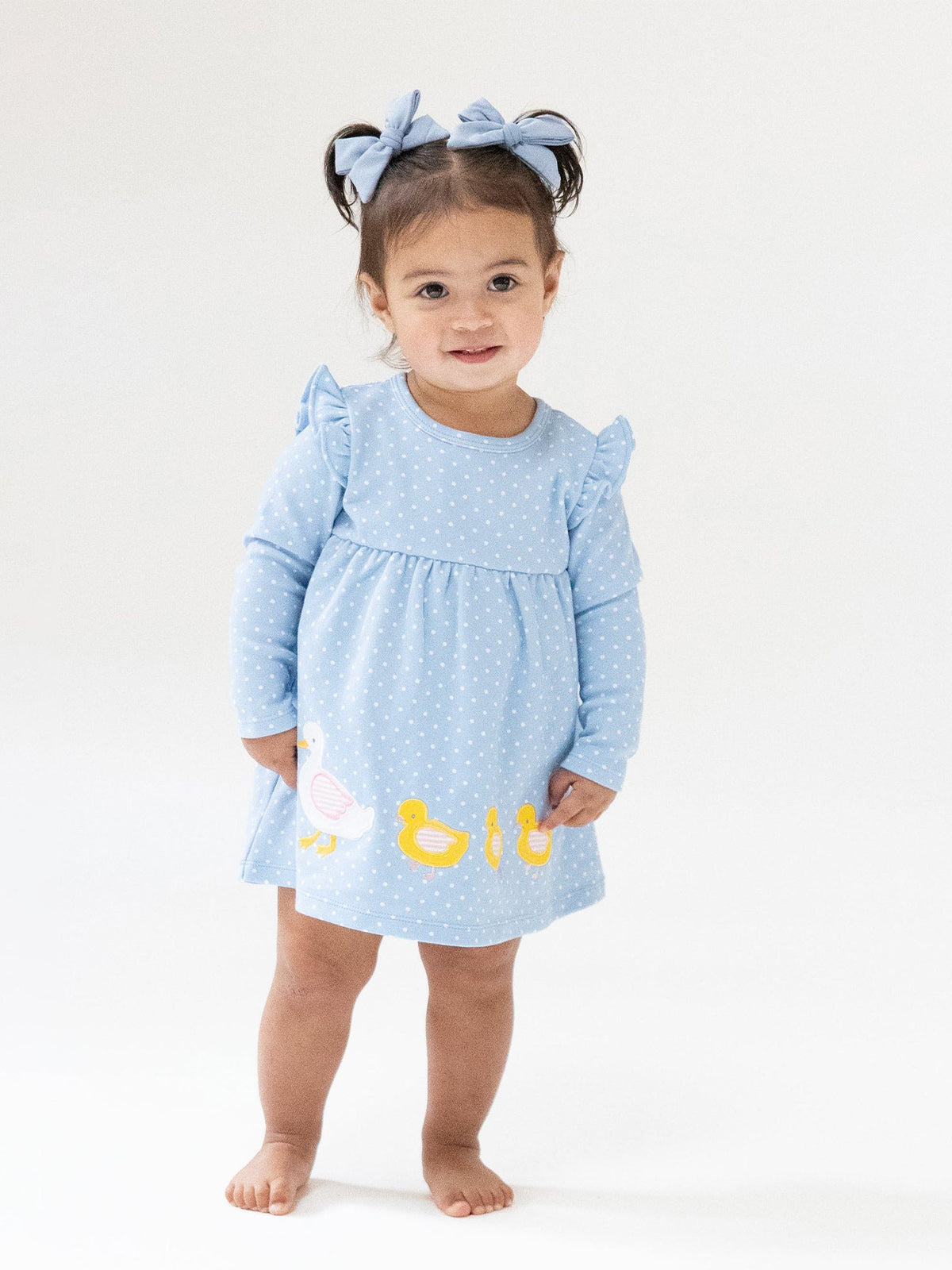 Toddler in a blue polka dot dress with duck designs, wearing bows in her hair, standing on a light background.