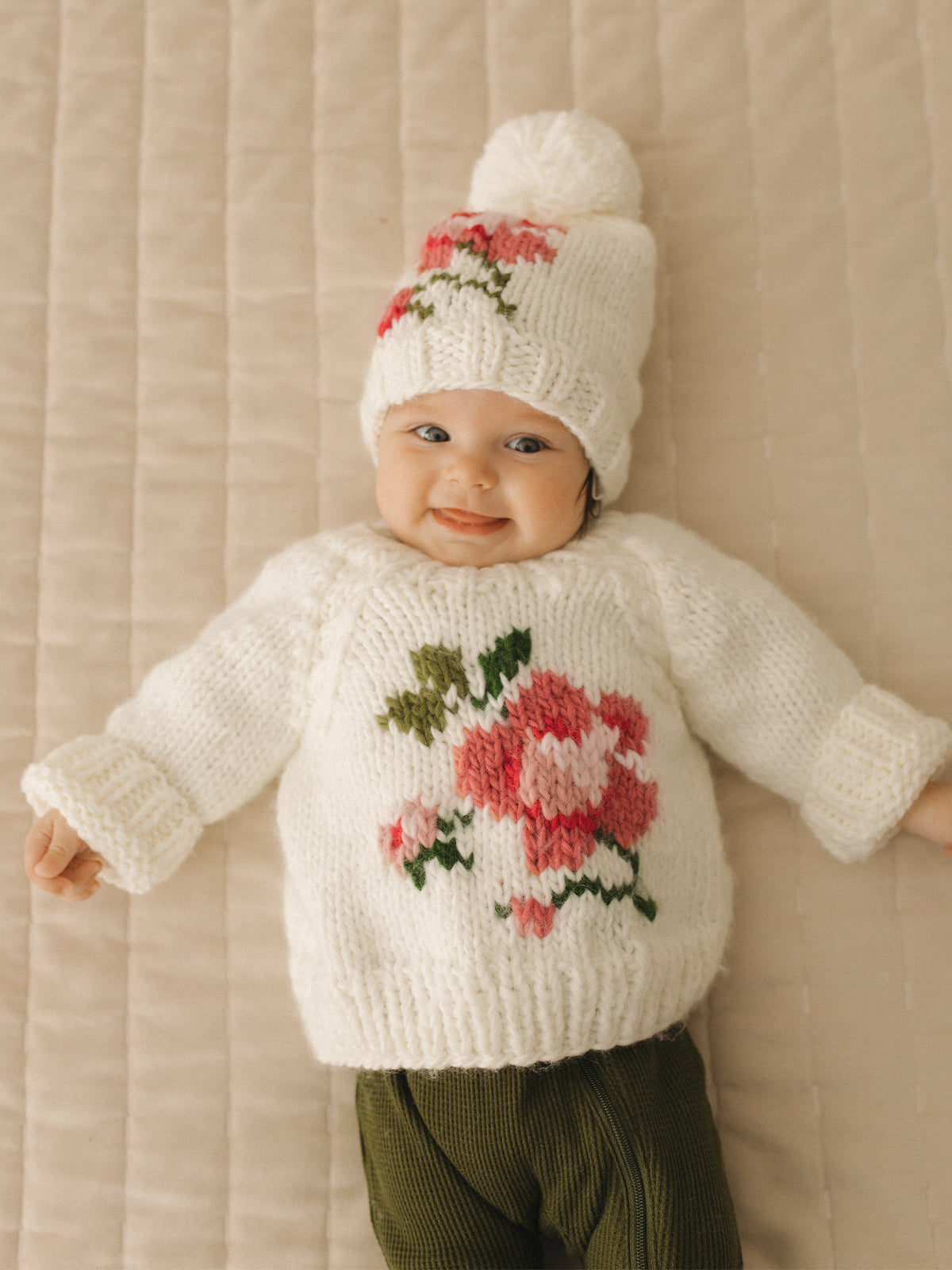 Smiling baby in a cozy knitted sweater with flower design and matching hat, lying on a textured blanket.