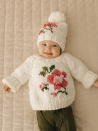 Smiling baby in a cozy knitted sweater with flower design and matching hat, lying on a textured blanket.