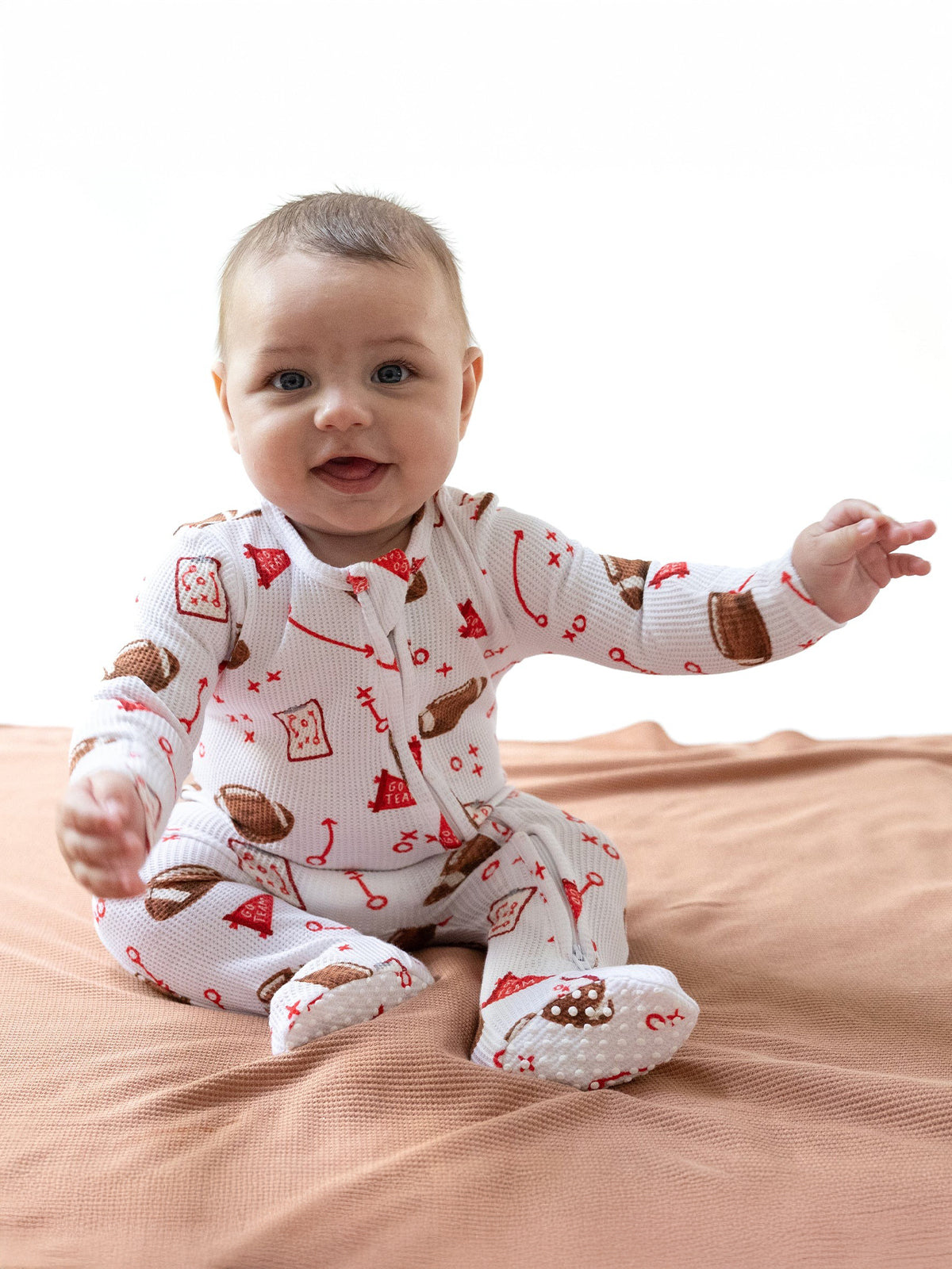 Smiling baby in patterned pajamas sits on a soft surface, reaching out with one hand.