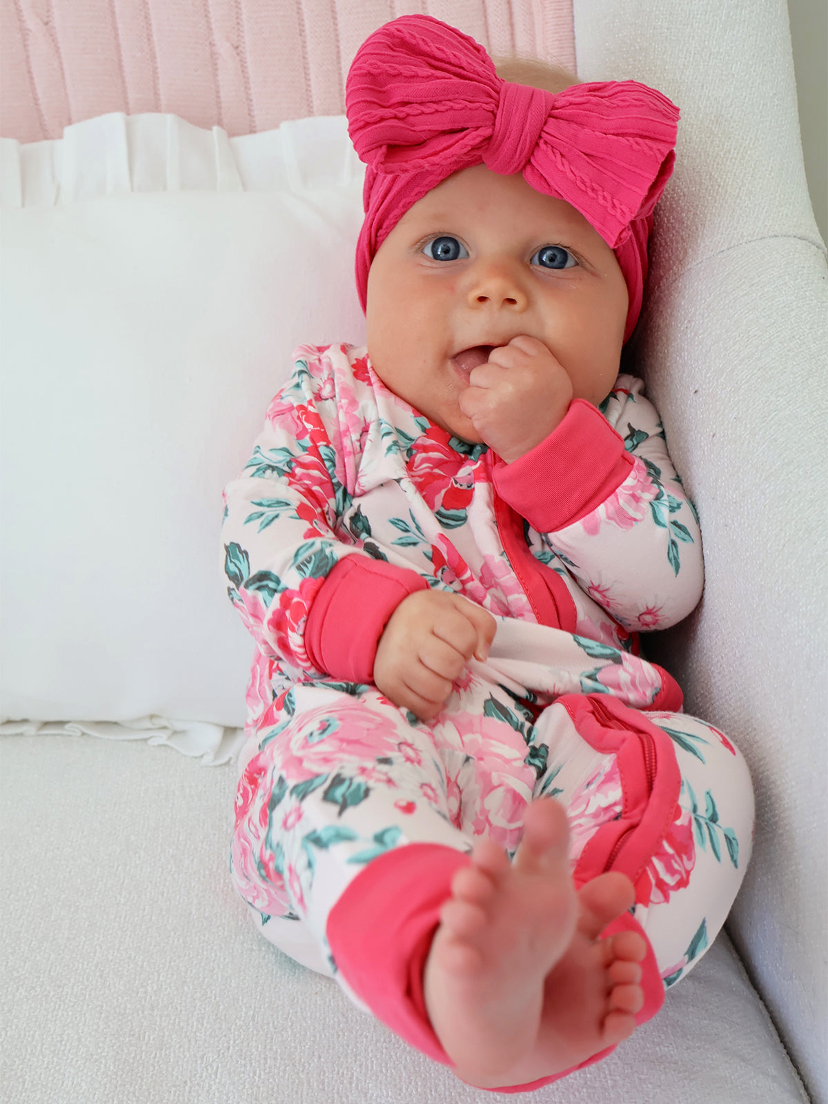 Smiling baby in floral pajamas with a pink headband, sitting on a couch, sucking thumb, playful expression.