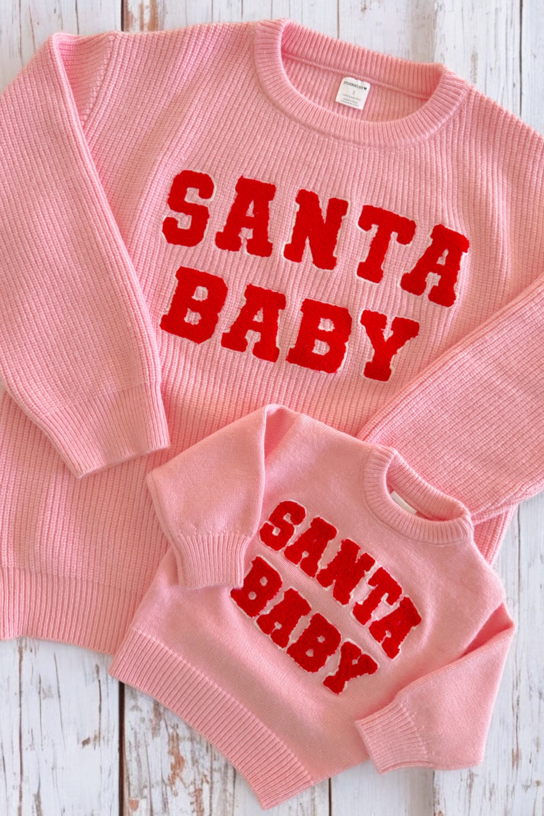 Pink "Santa Baby" sweaters in adult and child sizes laid flat on a wooden surface.