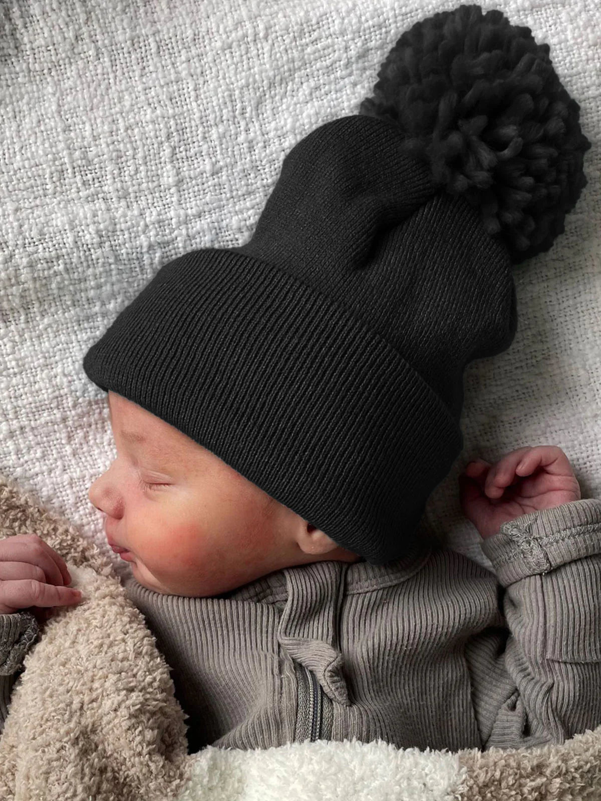 Newborn baby resting in a cozy blanket, wearing a black beanie with a pom-pom on top.