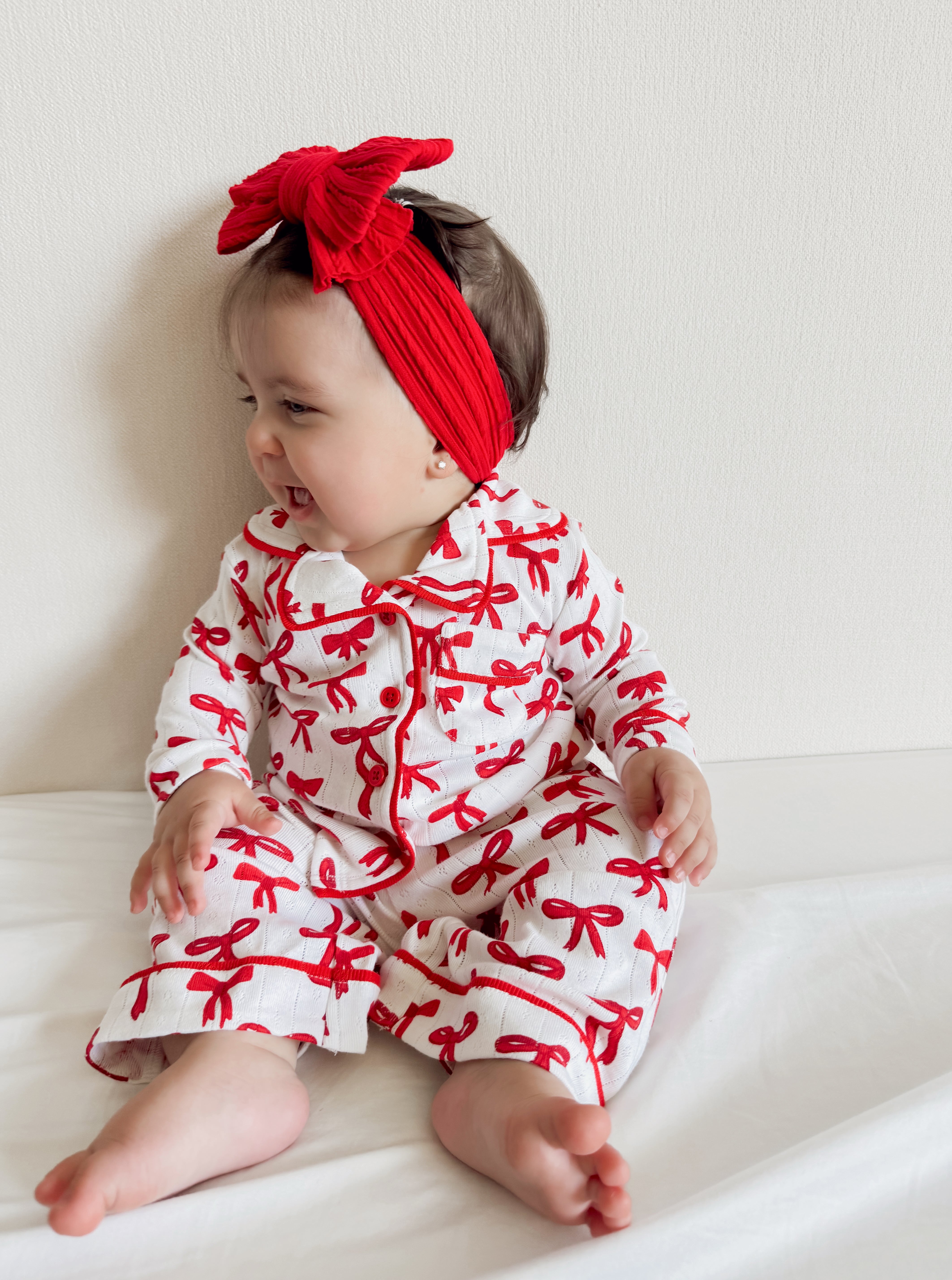 Smiling baby in a white outfit with red bows and a matching red headband, sitting on a white surface.