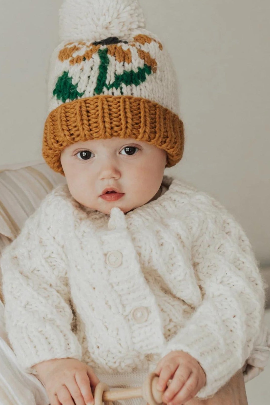 Baby wearing a cozy knitted sweater and colorful pom-pom hat, holding a wooden toy while looking at the camera.