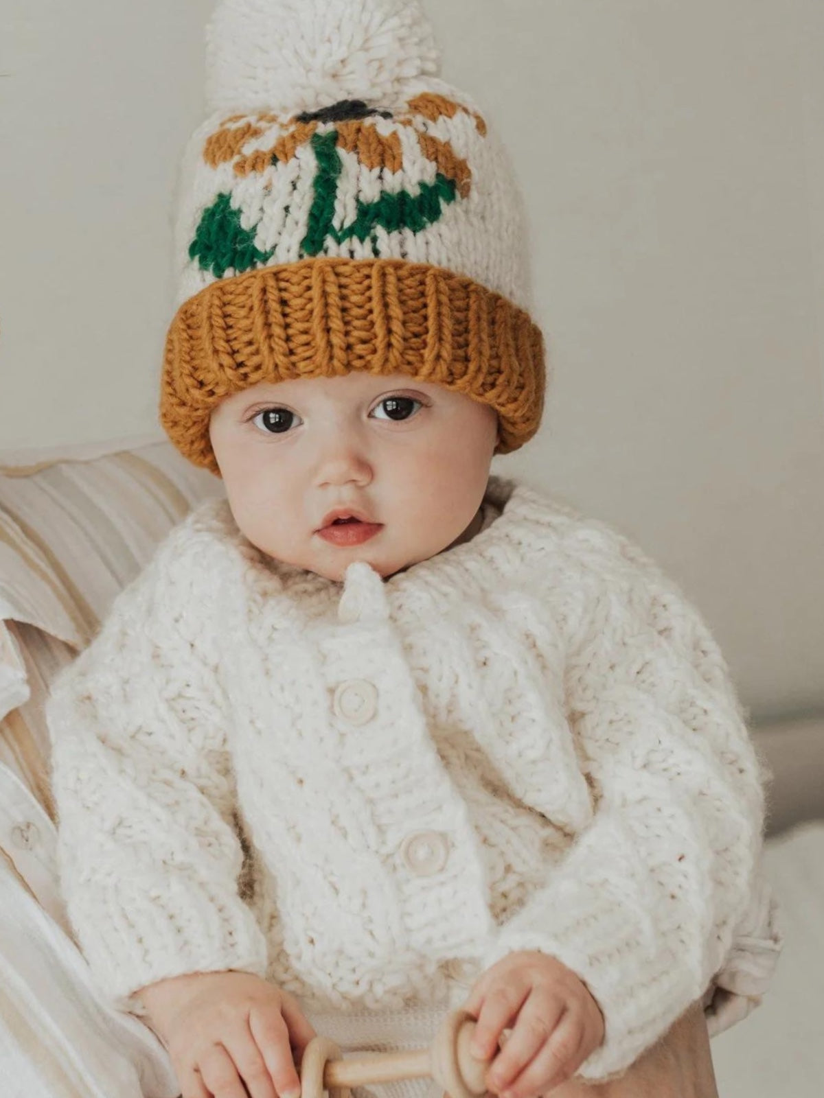 Baby wearing a cozy knitted sweater and colorful pom-pom hat, holding a wooden toy while looking at the camera.