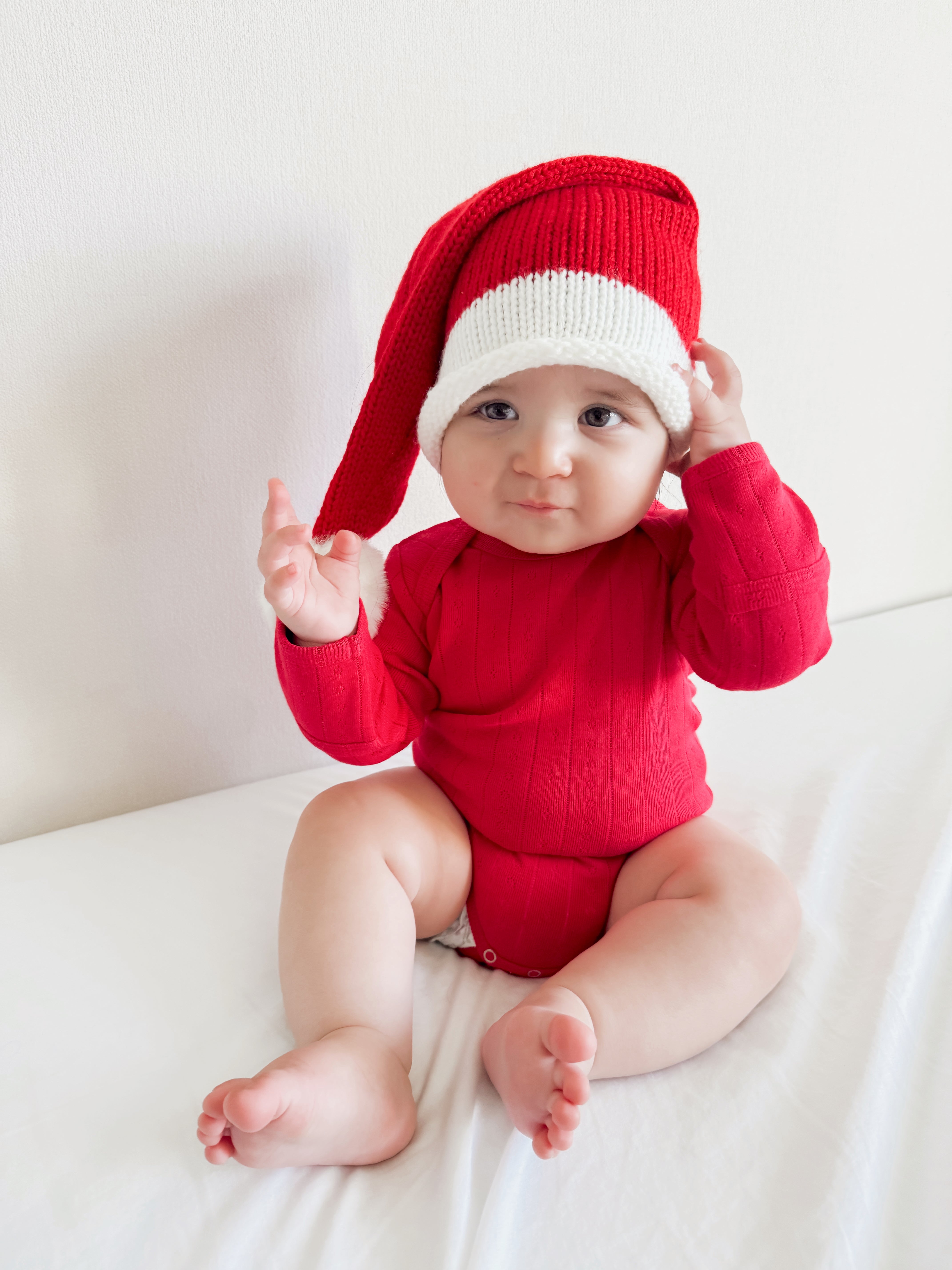 Baby in red outfit and festive Santa hat, sitting on a white surface, looking at the camera.