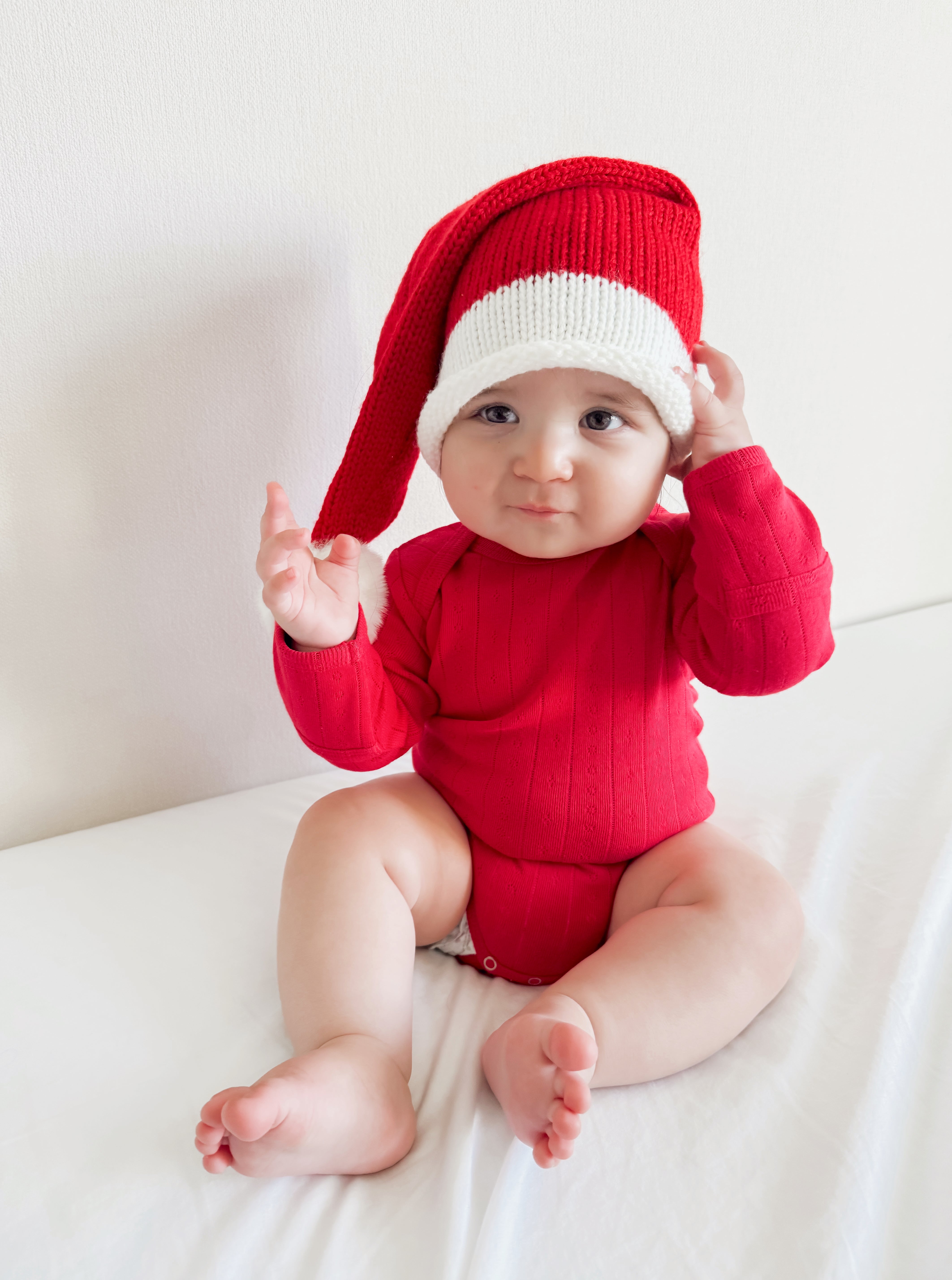 Baby in red outfit and festive Santa hat, sitting on a white surface, looking at the camera.