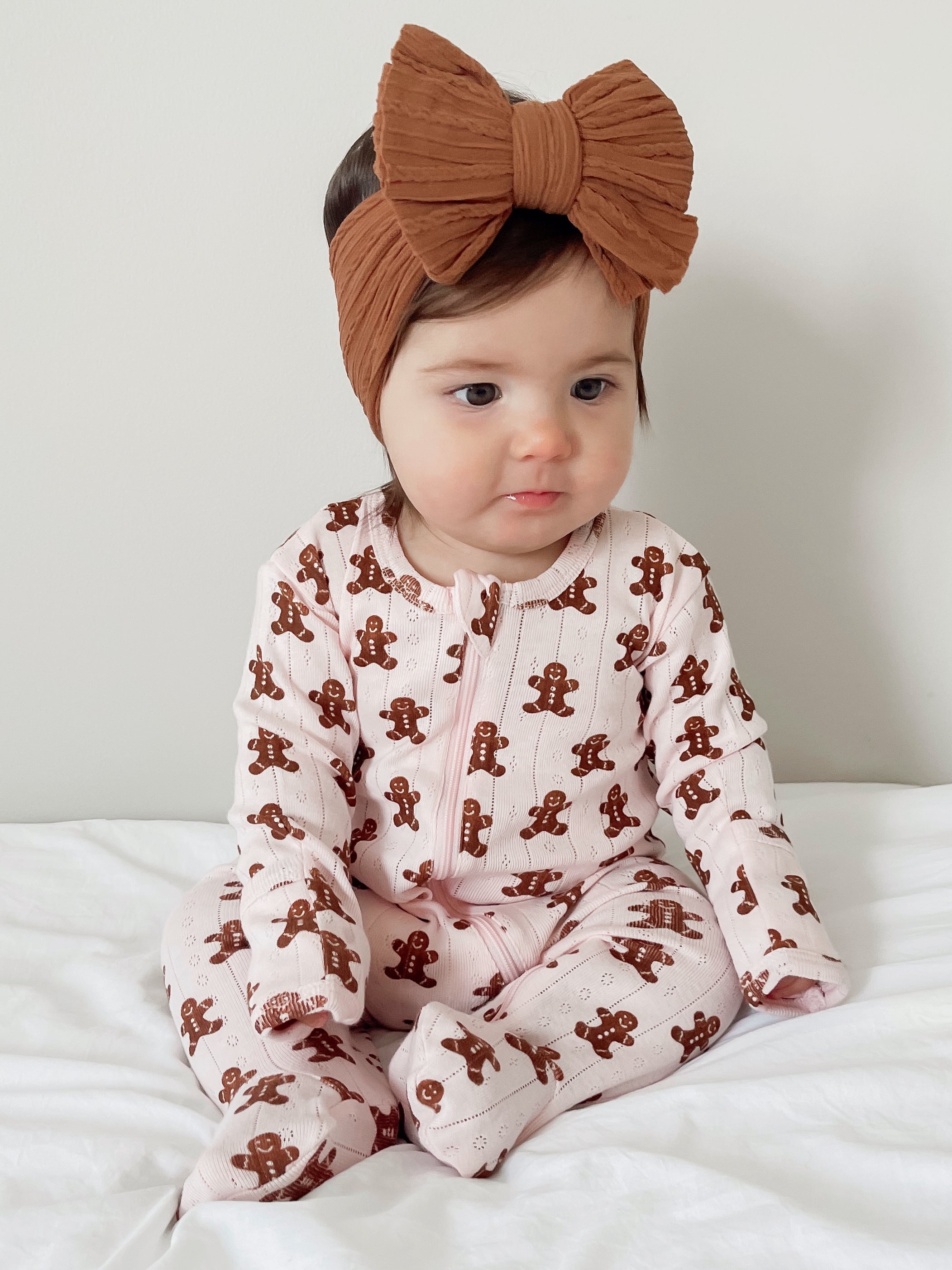 Baby in gingerbread-patterned pajamas with a large brown bow headband, seated on a white surface.