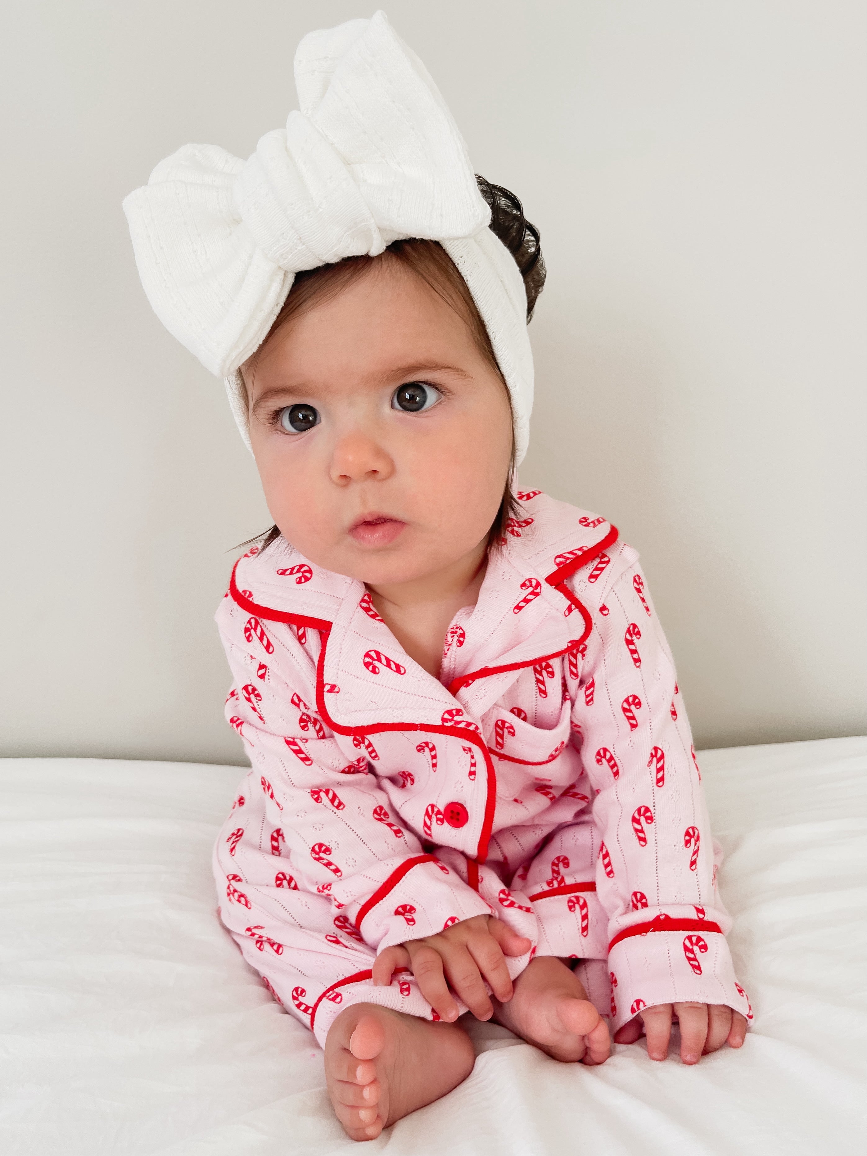Baby wearing pink candy cane pajamas and a white bow headband, sitting on a bed with a neutral background.