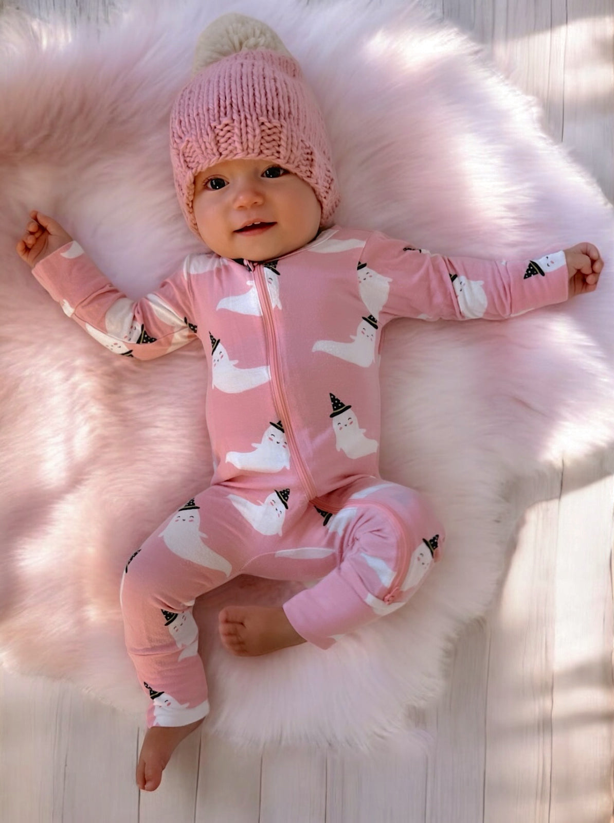 Baby lying on a fluffy rug, wearing a pink onesie with ghost prints and a knitted hat, smiling at the camera.