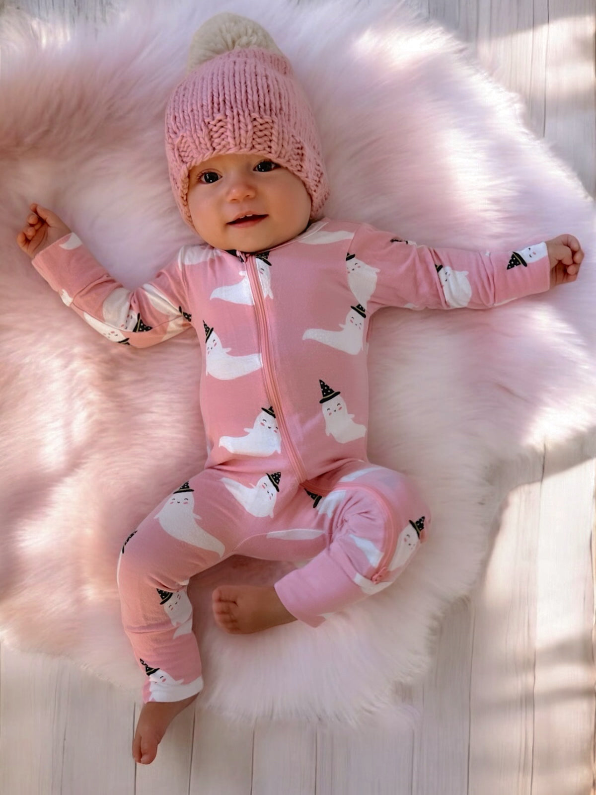 Baby lying on a fluffy rug, wearing a pink onesie with ghost prints and a knitted hat, smiling at the camera.