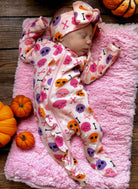 Sleeping baby in a colorful skull-patterned outfit on a pink blanket, surrounded by small pumpkins.