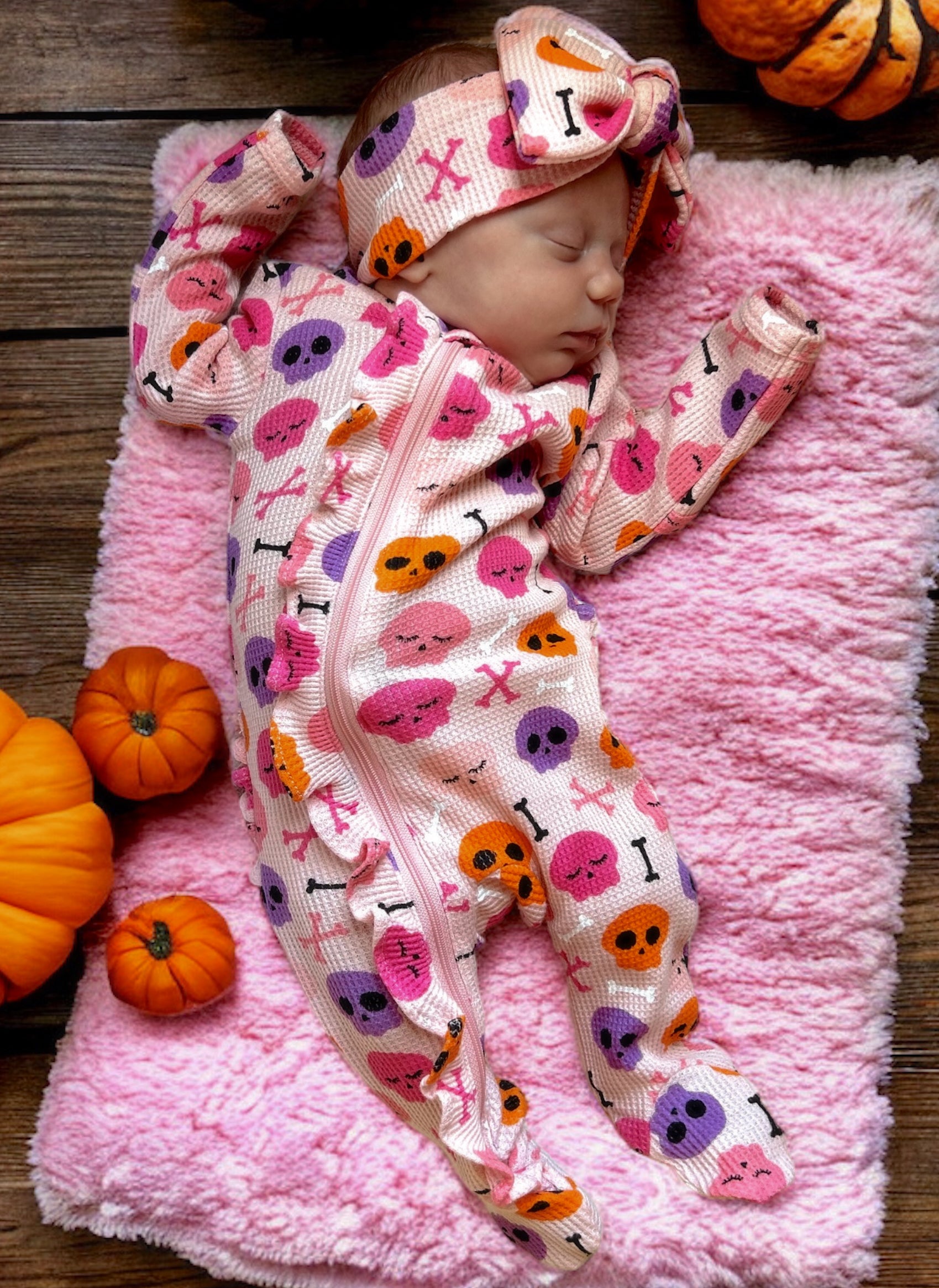 Sleeping baby in a colorful skull-patterned outfit on a pink blanket, surrounded by small pumpkins.