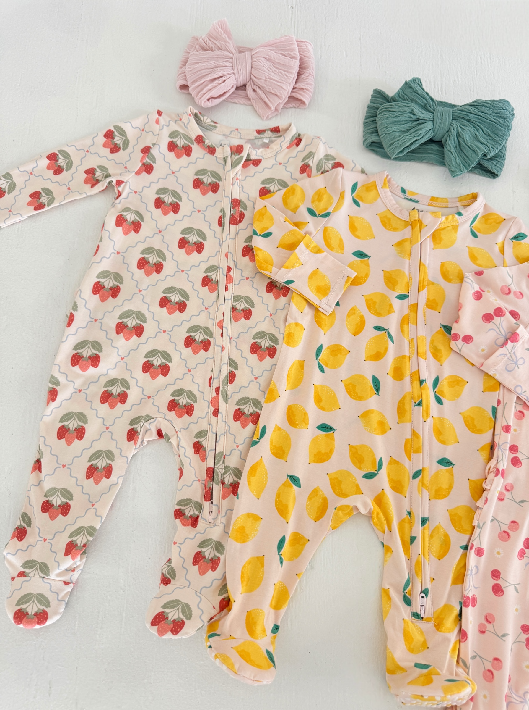 Two infant onesies with strawberry and lemon patterns, accompanied by pink and green fabric headbands.