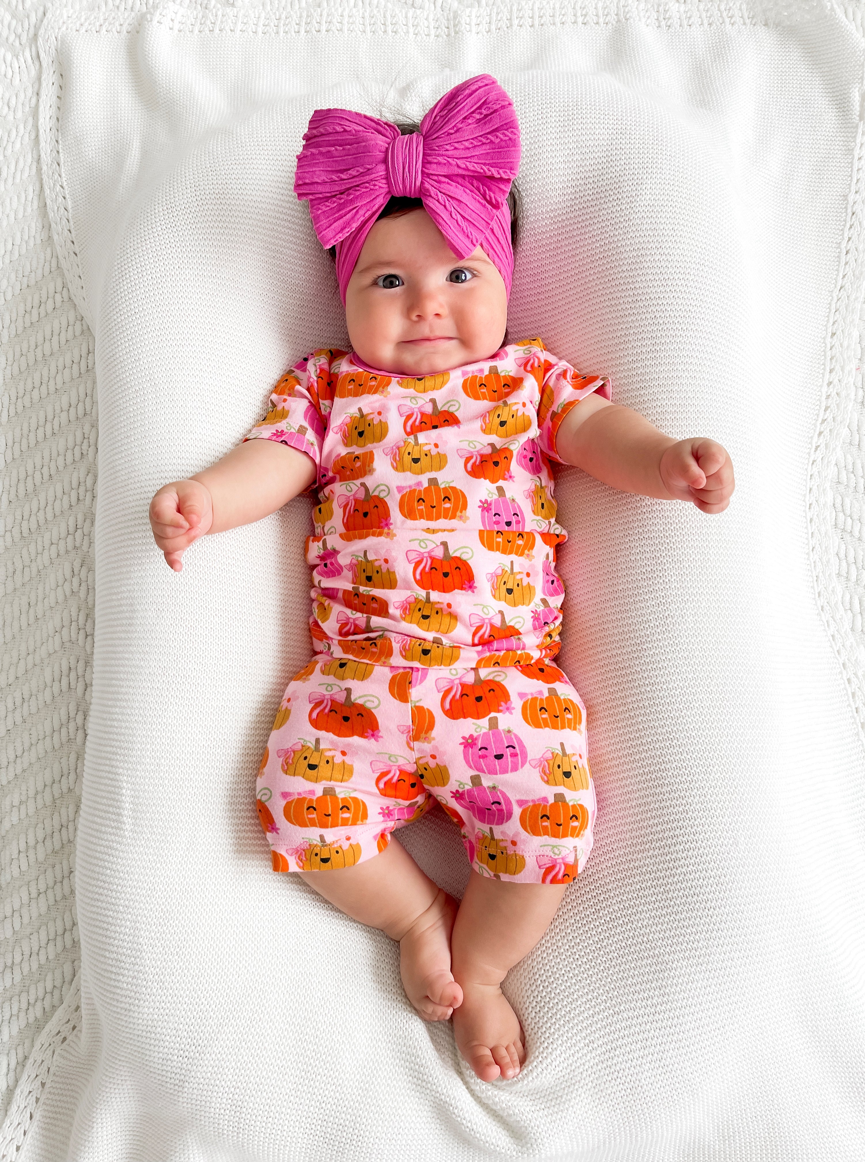 Infant with a large pink bow, wearing a pumpkin-patterned outfit, lying on a white blanket.