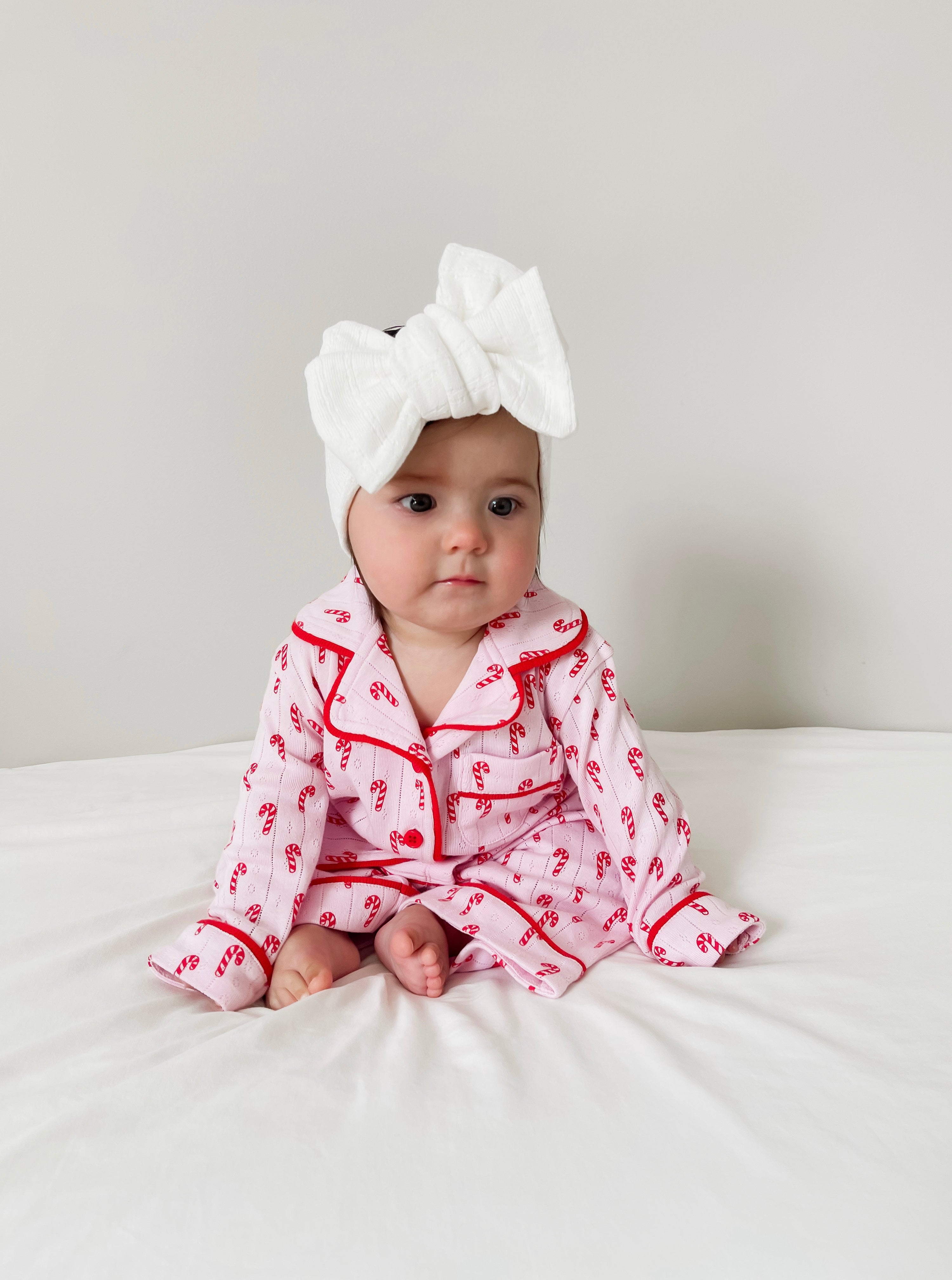 A baby sits on a white bed wearing pink pajamas with candy cane patterns and a large white bow headband.