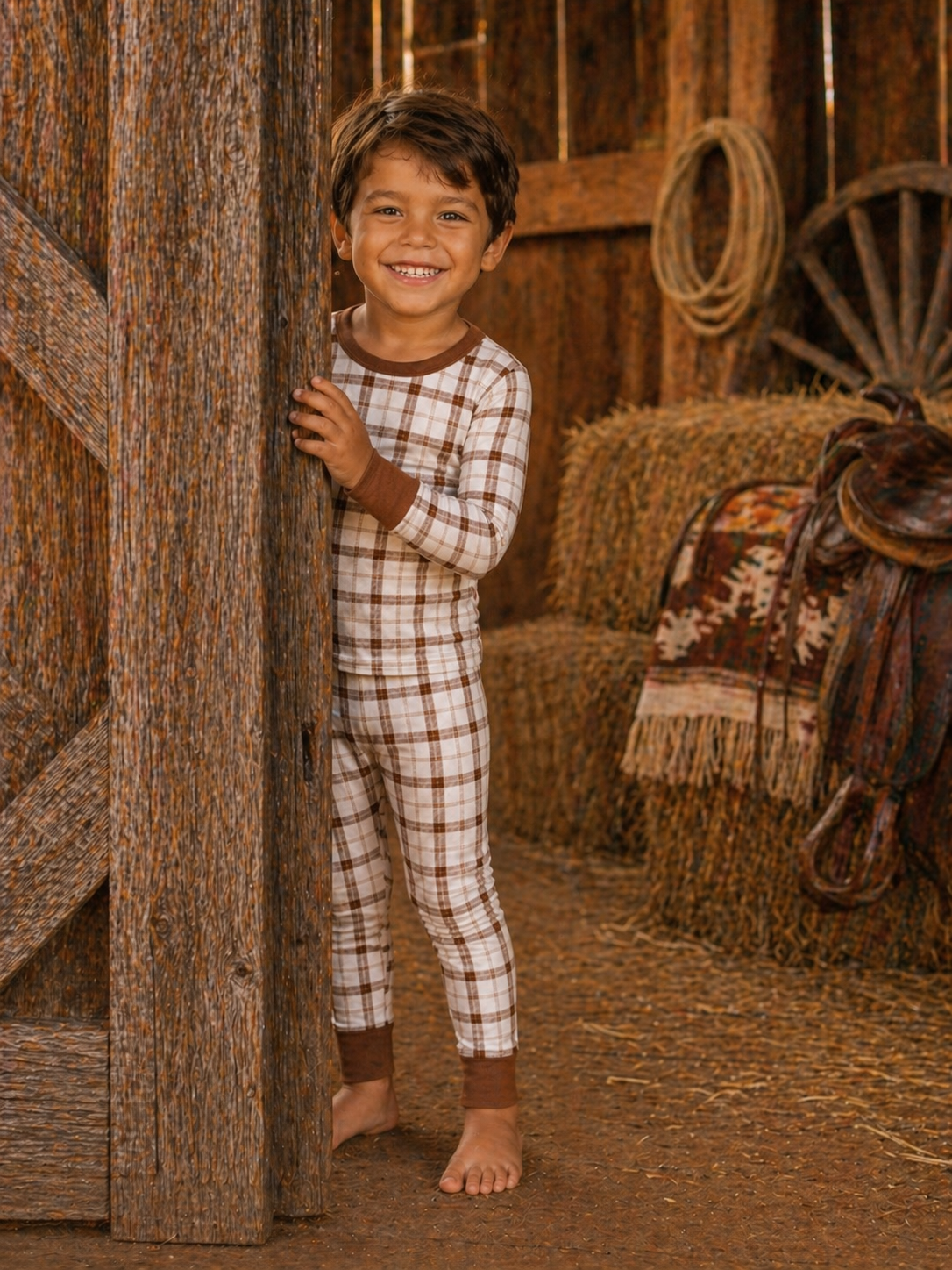 Smiling child in plaid pajamas peeking from a barn door, surrounded by hay bales and rustic decor.