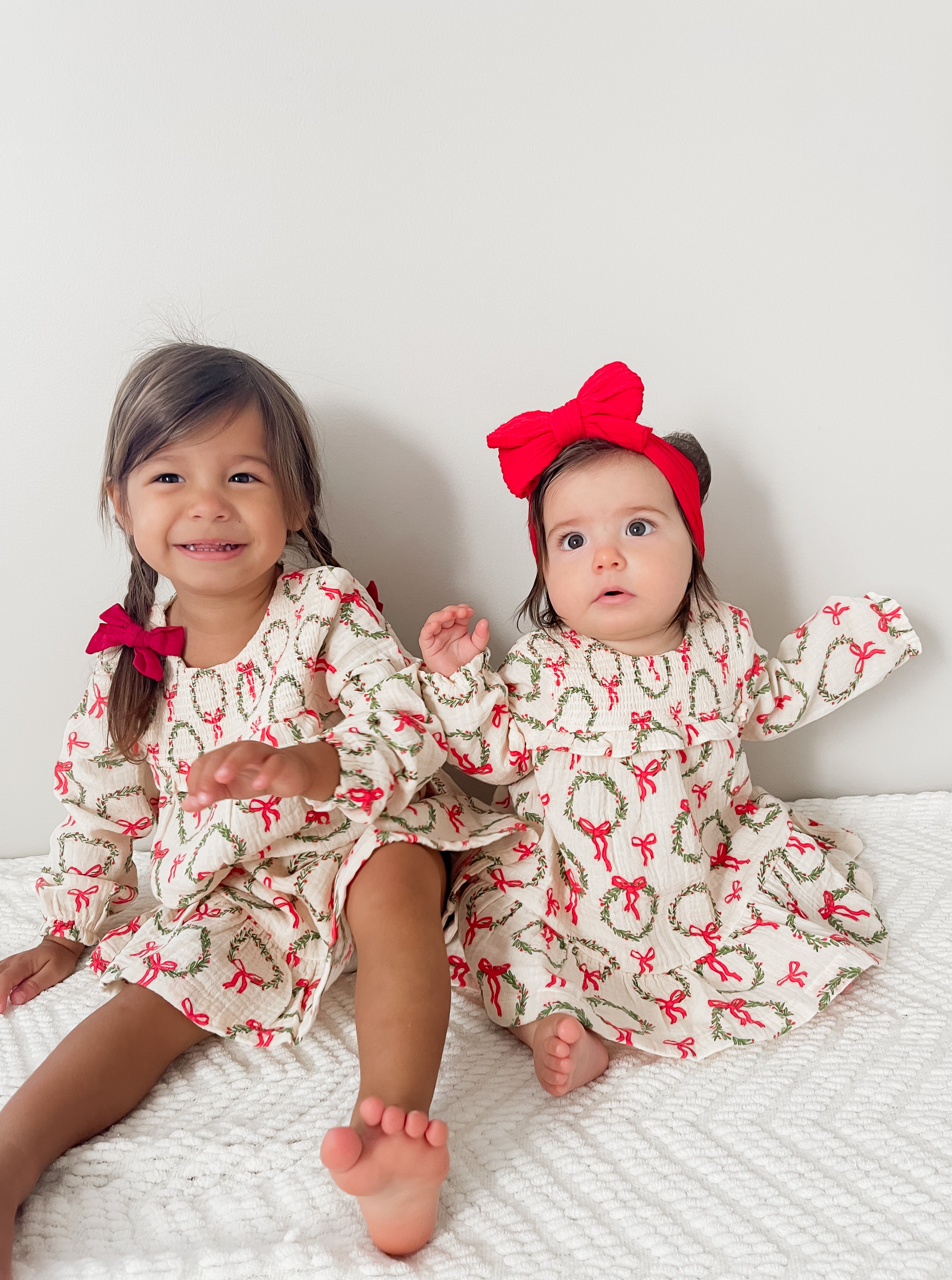 Two young girls in festive dresses with bows, sitting on a white blanket against a plain background.