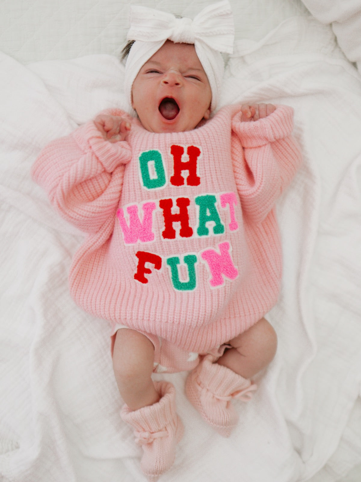 Baby yawning in a pink sweater with "OH WHAT FUN" text, wearing a headband and cozy booties, on a white blanket.