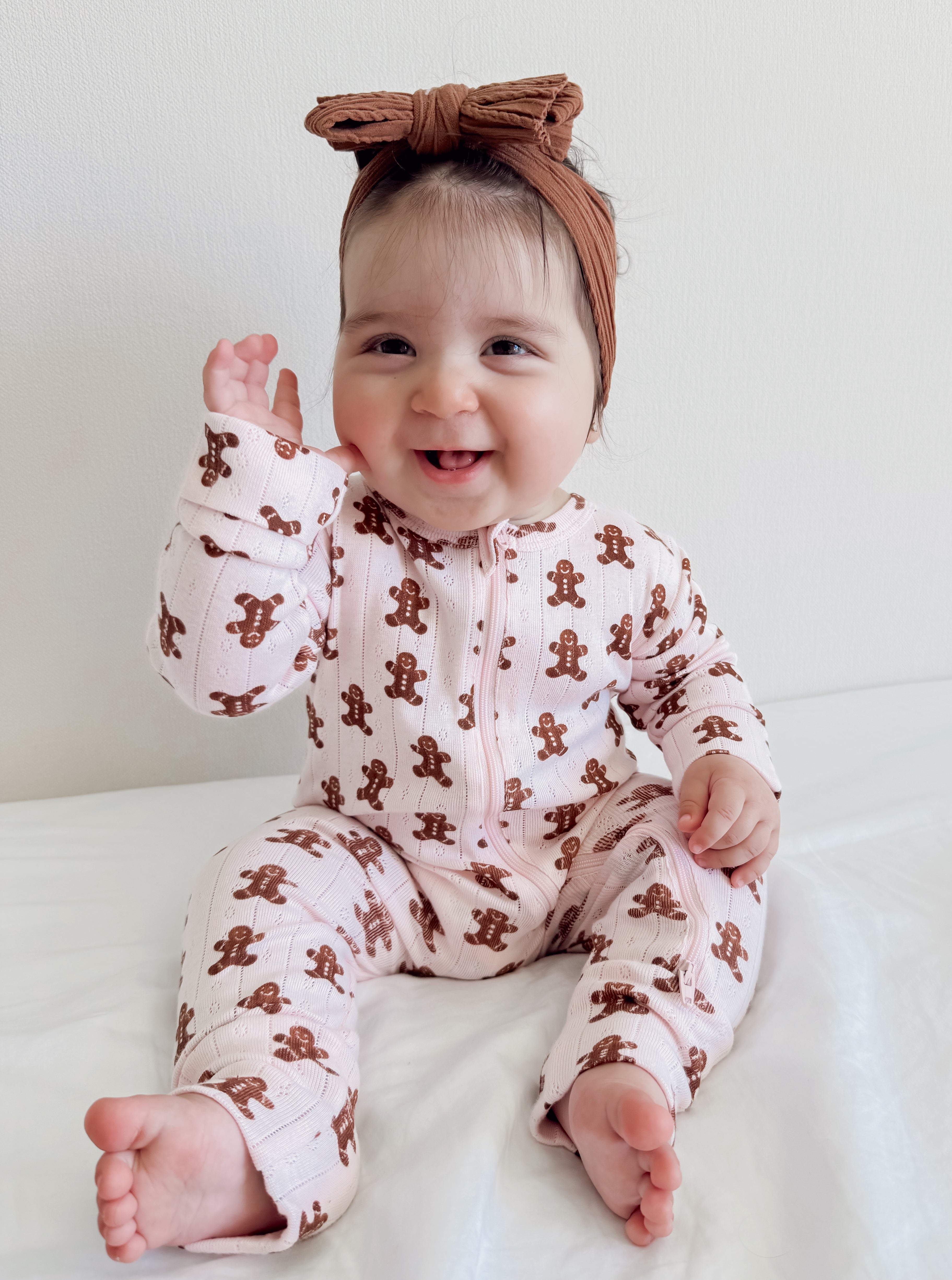 Smiling baby in pink gingerbread-patterned onesie and brown headband waving on a white background.