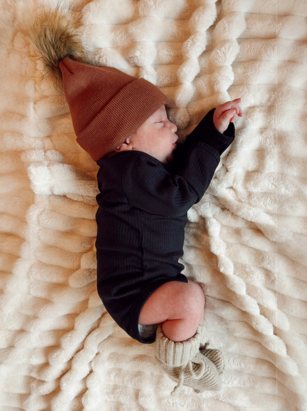 Newborn baby sleeping on a fluffy blanket, wearing a brown hat and knitted booties.