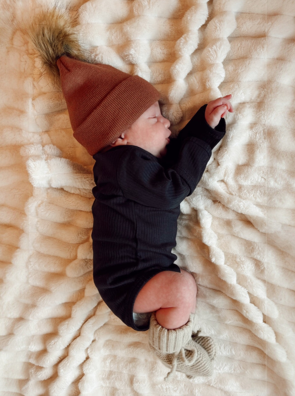 Newborn baby sleeping on a fluffy blanket, wearing a brown hat and knitted booties.