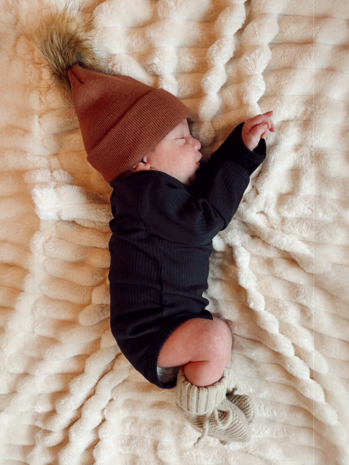 Newborn baby sleeping on a fluffy blanket, wearing a brown hat and knitted booties.