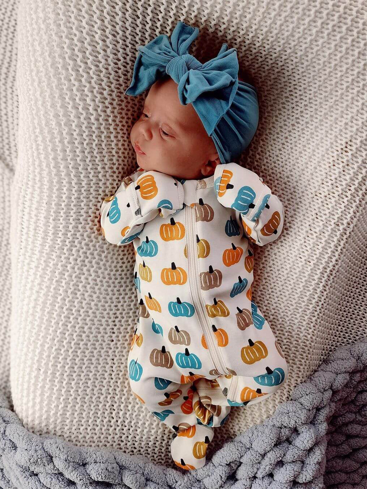 Baby dressed in a pumpkin-patterned onesie, wearing a teal bow headband, resting on a cozy blanket.