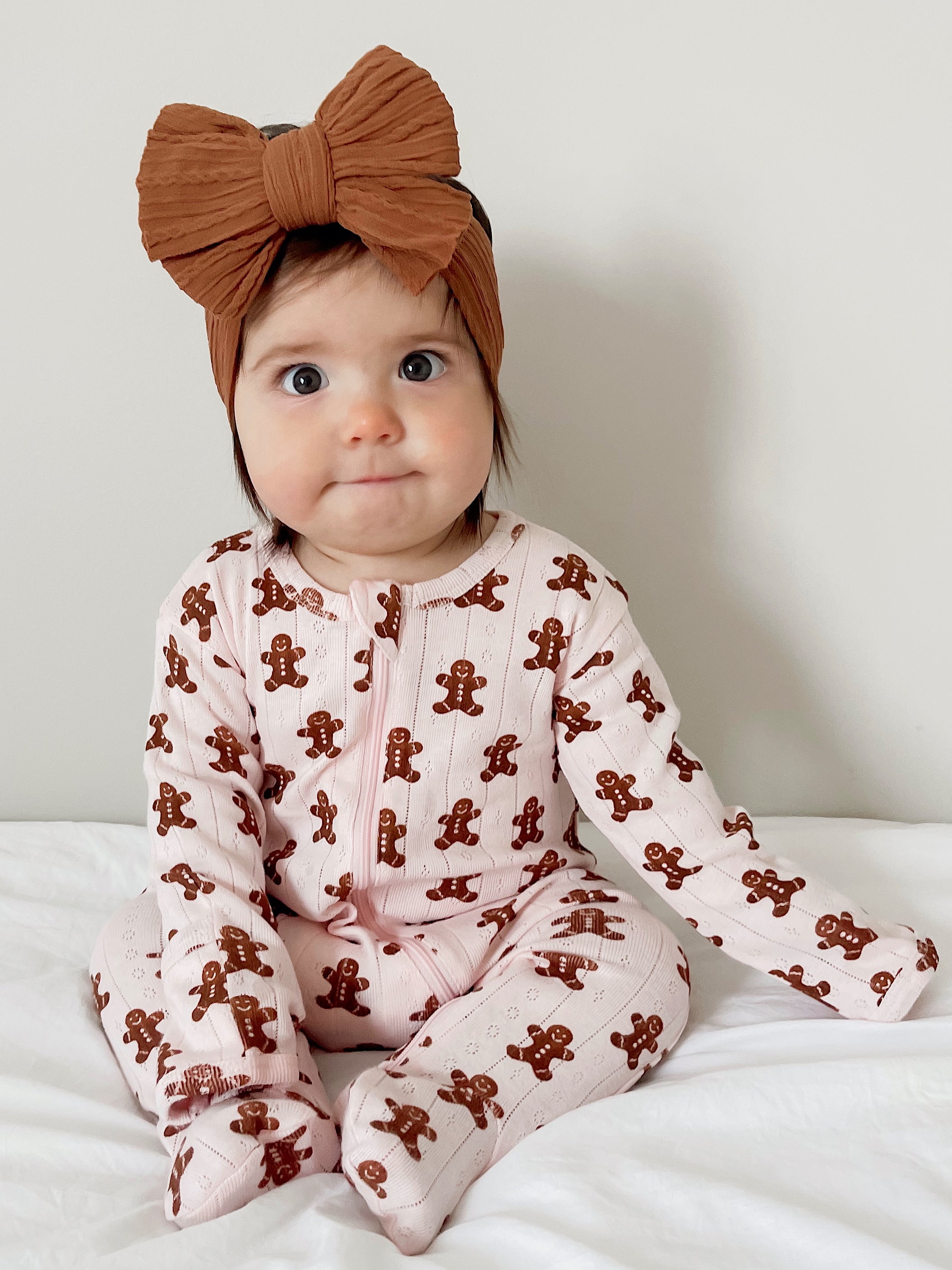 Baby wearing a pink gingerbread-patterned onesie and a large brown headband, sitting on a white blanket.