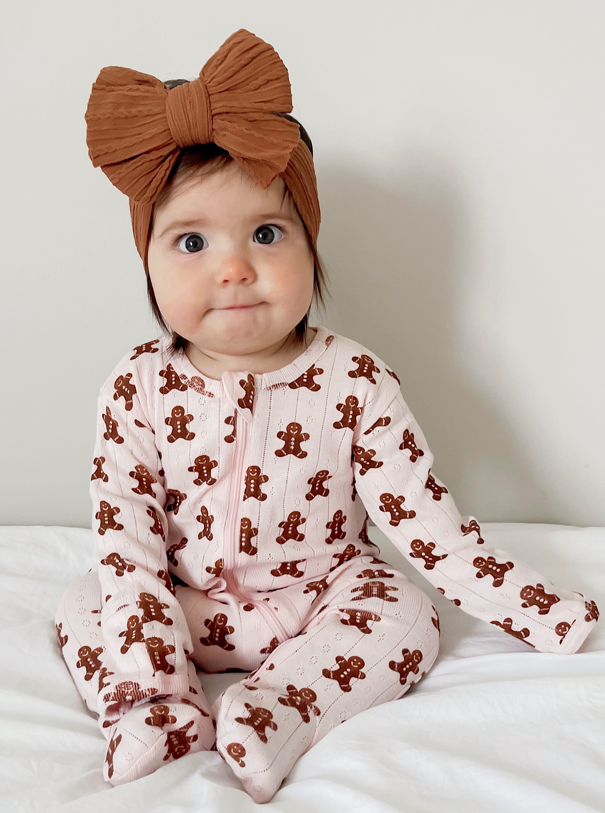 Baby wearing a pink gingerbread-patterned onesie and a large brown headband, sitting on a white blanket.