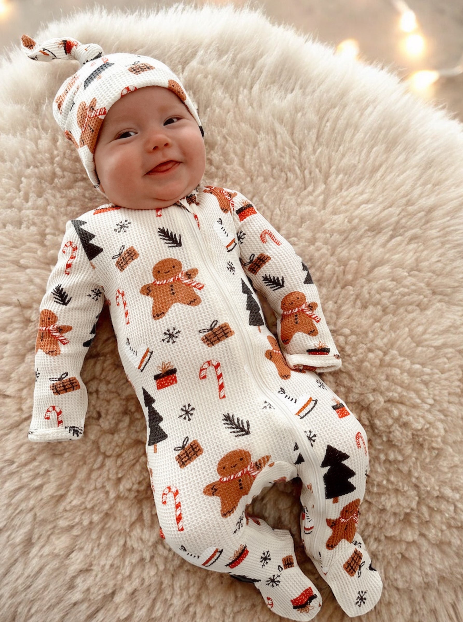 Baby in a festive gingerbread-patterned pajamas and hat, smiling while lying on a fluffy surface.