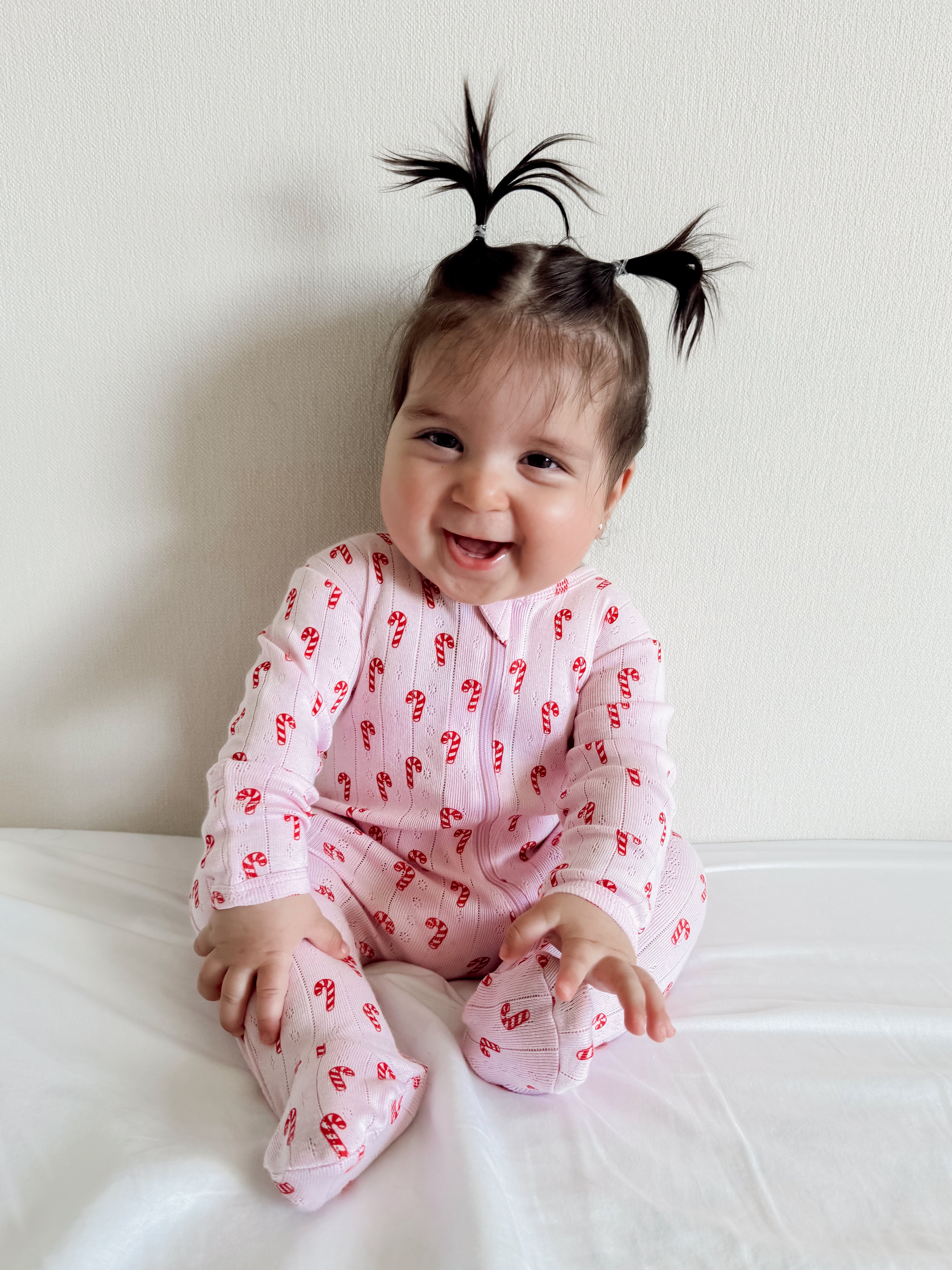 Smiling baby in pink pajamas with candy cane patterns, sitting on a white surface against a light wall.