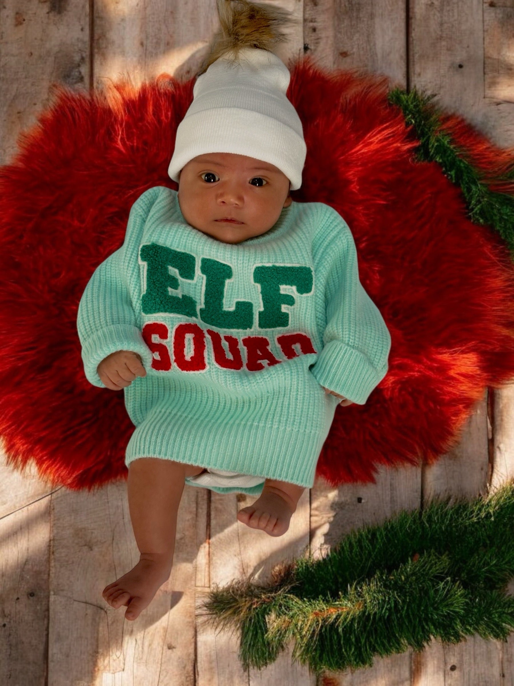 Baby in a mint green sweater with "ELF SQUAD" text, lying on a red plush round mat with greenery around.