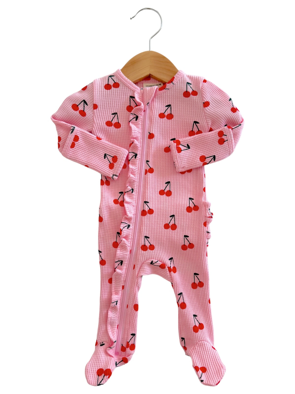 Light pink baby onesie with cherry prints, featuring a front zipper and ruffle details on the side. Hanging on a wooden hanger.