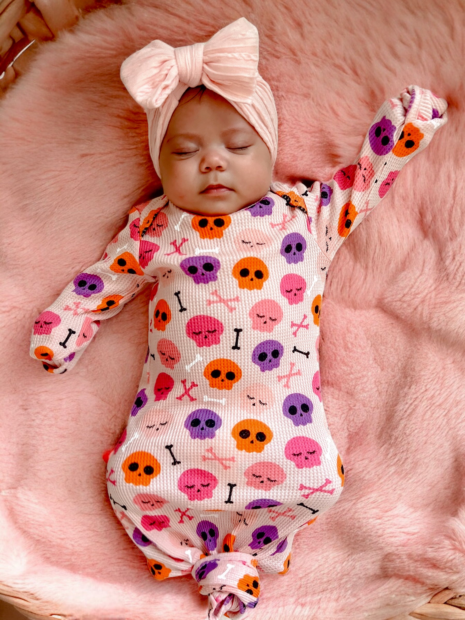 Baby sleeping on a pink blanket, wearing a colorful skull-print outfit and a large bow headband.
