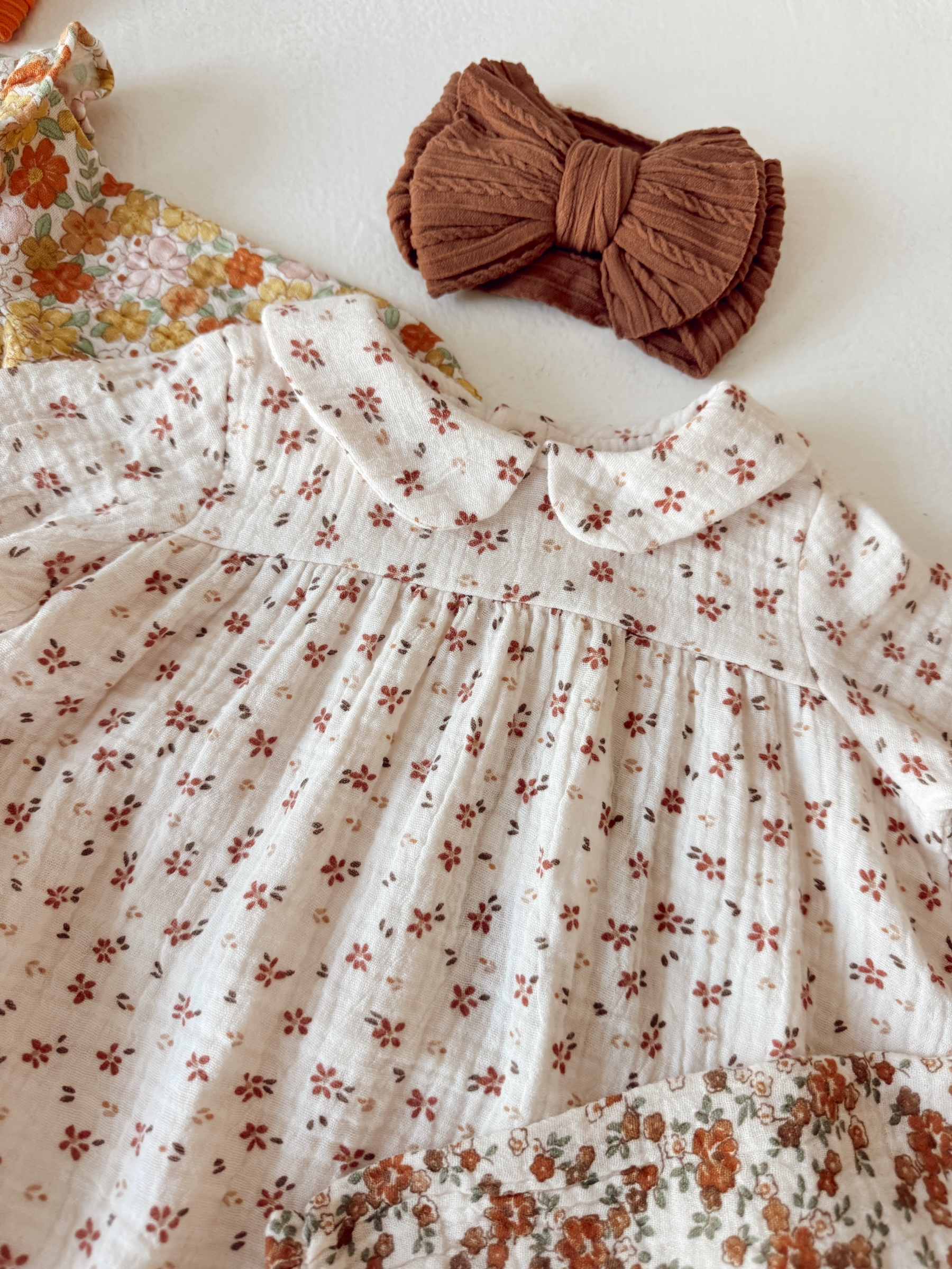 Floral baby dress with a collar and a brown fabric bow hair accessory on a light surface.