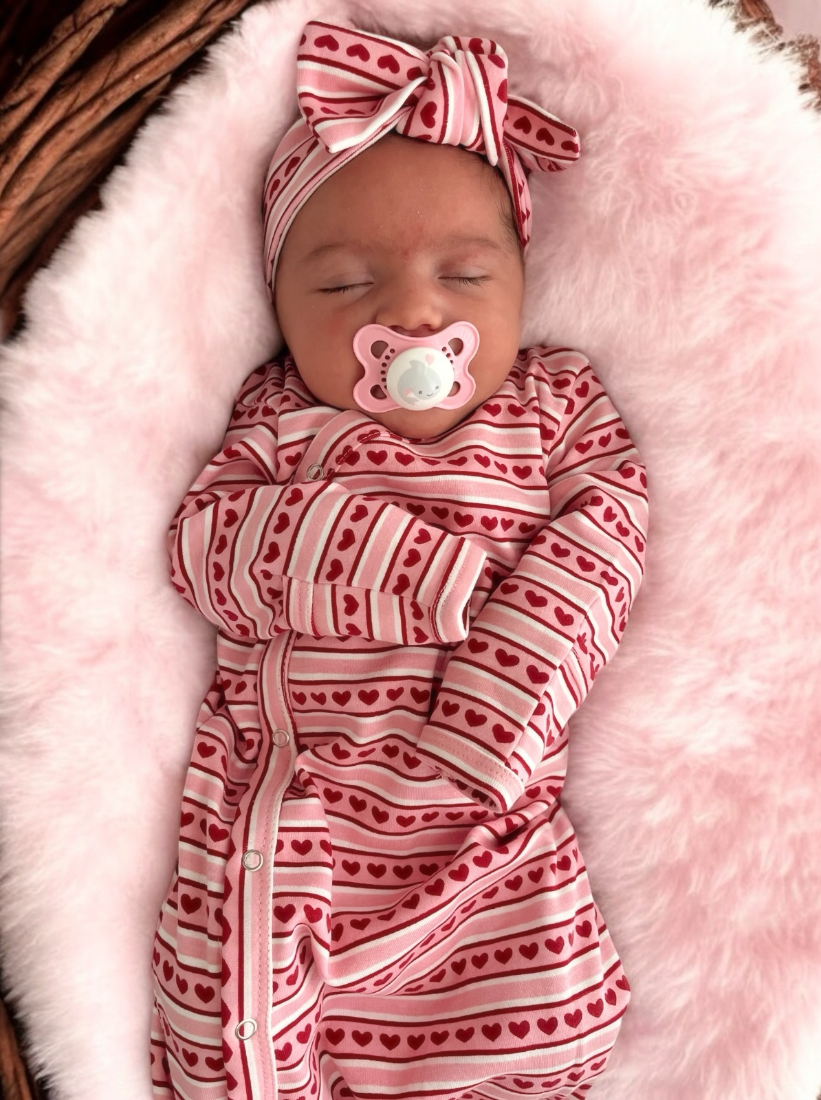 Sleeping baby in heart-patterned outfit with a pink pacifier, nestled on a soft, fluffy pink blanket.