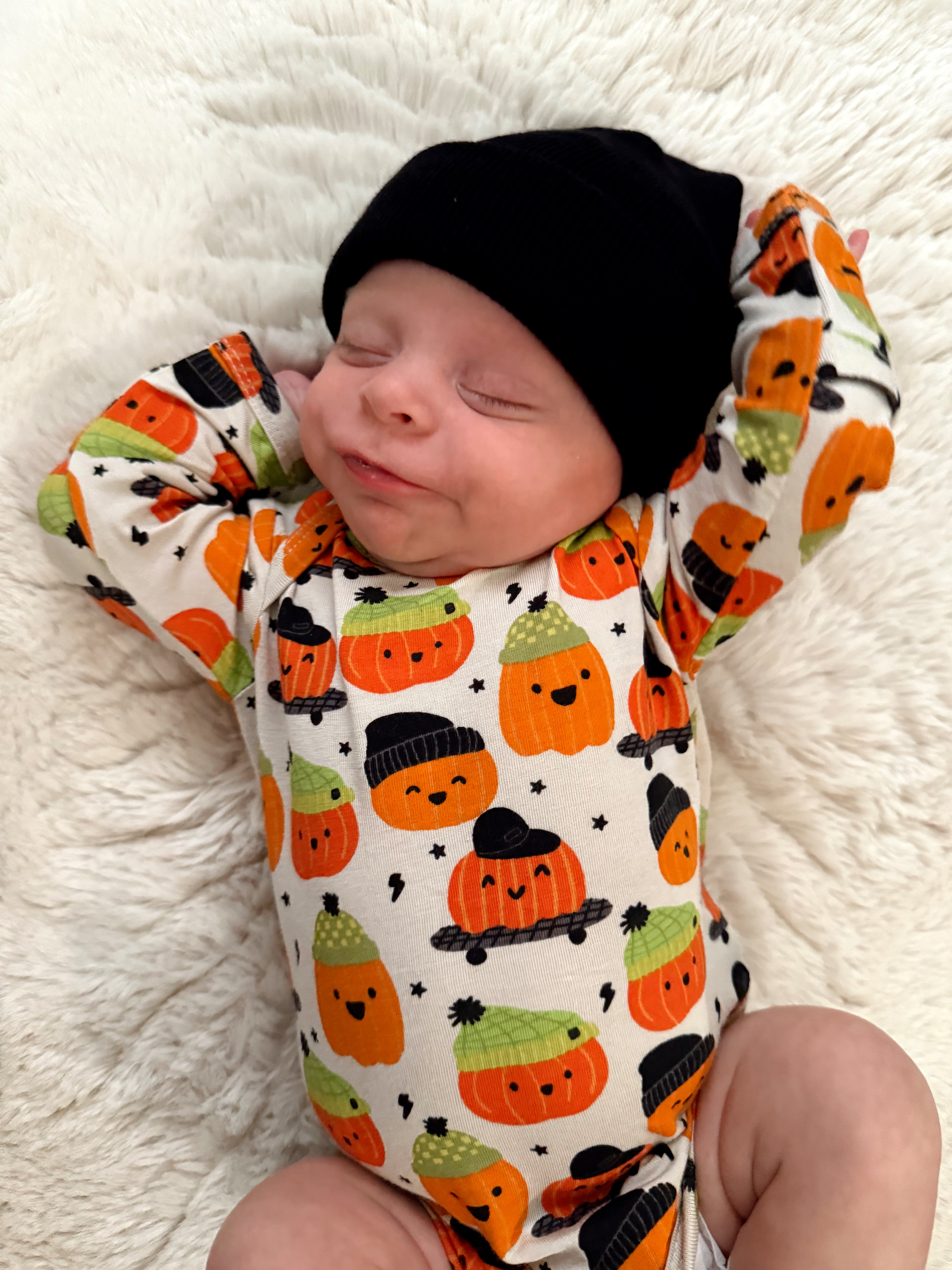 Smiling baby in a cute pumpkin-patterned outfit and black beanie, resting comfortably on a fluffy white blanket.