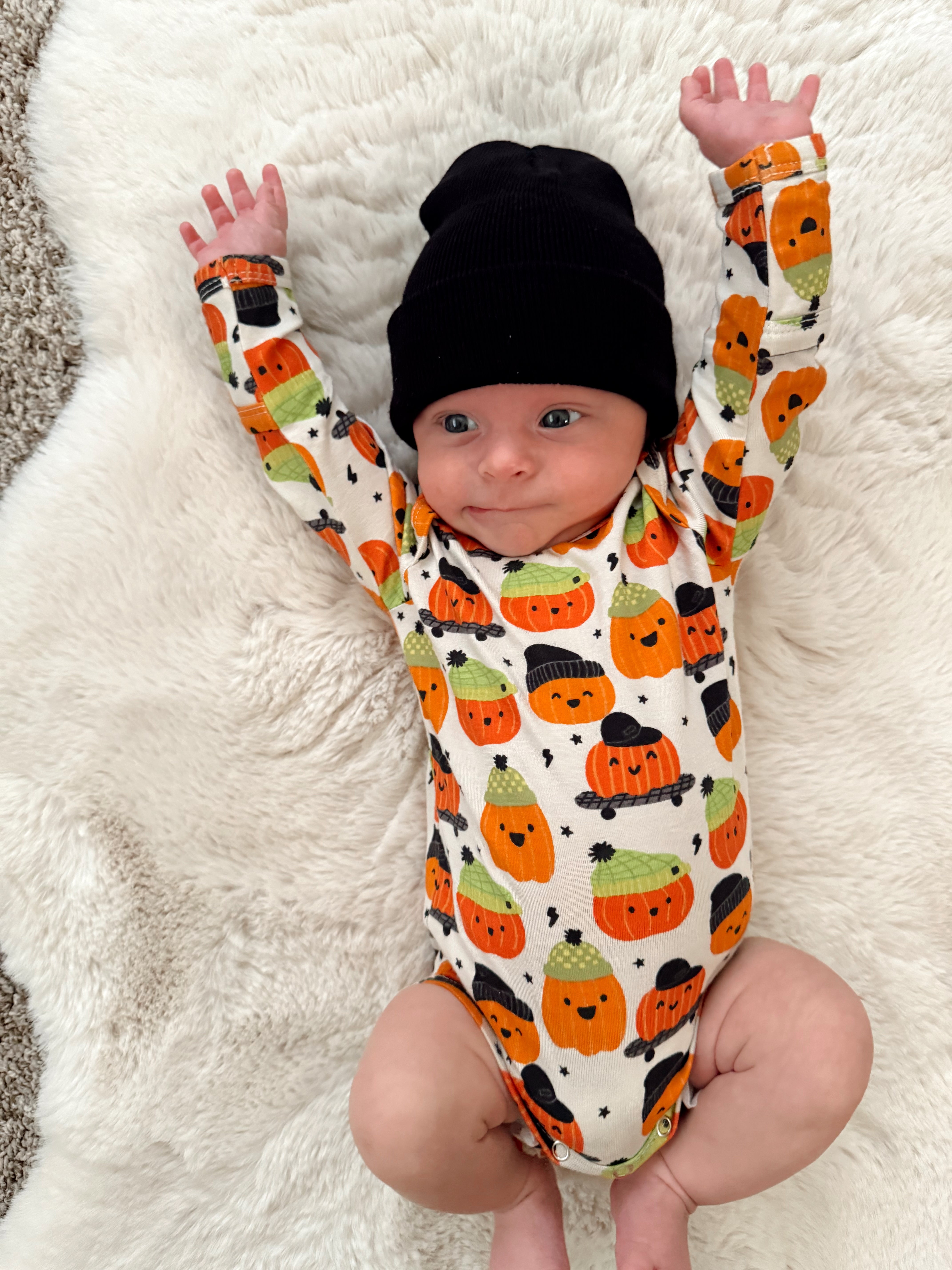 Infant wearing a pumpkin-patterned onesie and a black beanie, joyfully lying on a fluffy white blanket.