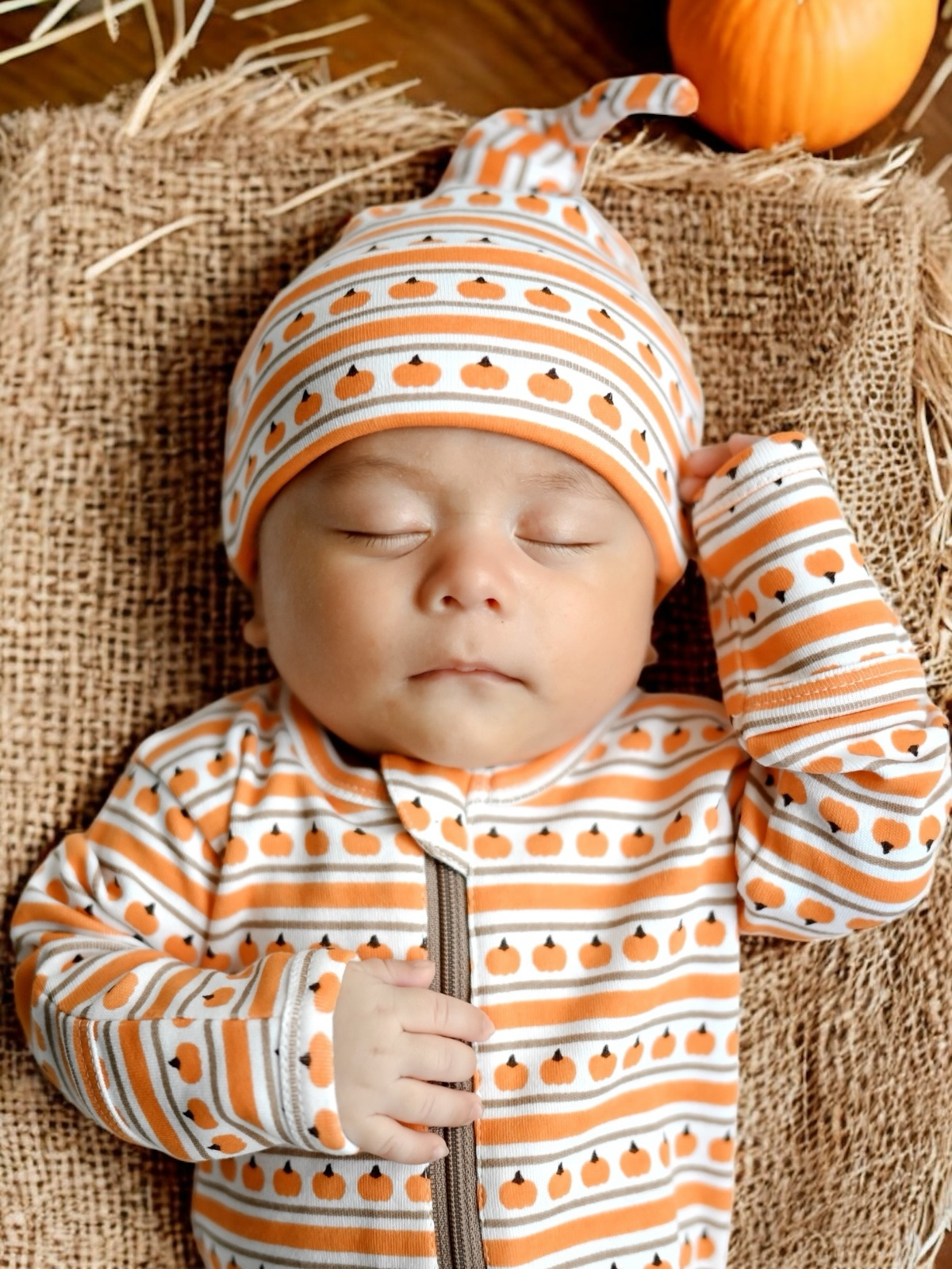 Sleeping baby in pumpkin-themed outfit on a burlap backdrop, with a pumpkin nearby.