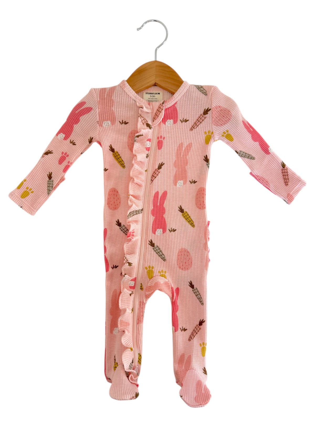 Pink baby romper with bunny and carrot patterns, featuring a ruffled front and long sleeves, hung on a wooden hanger.