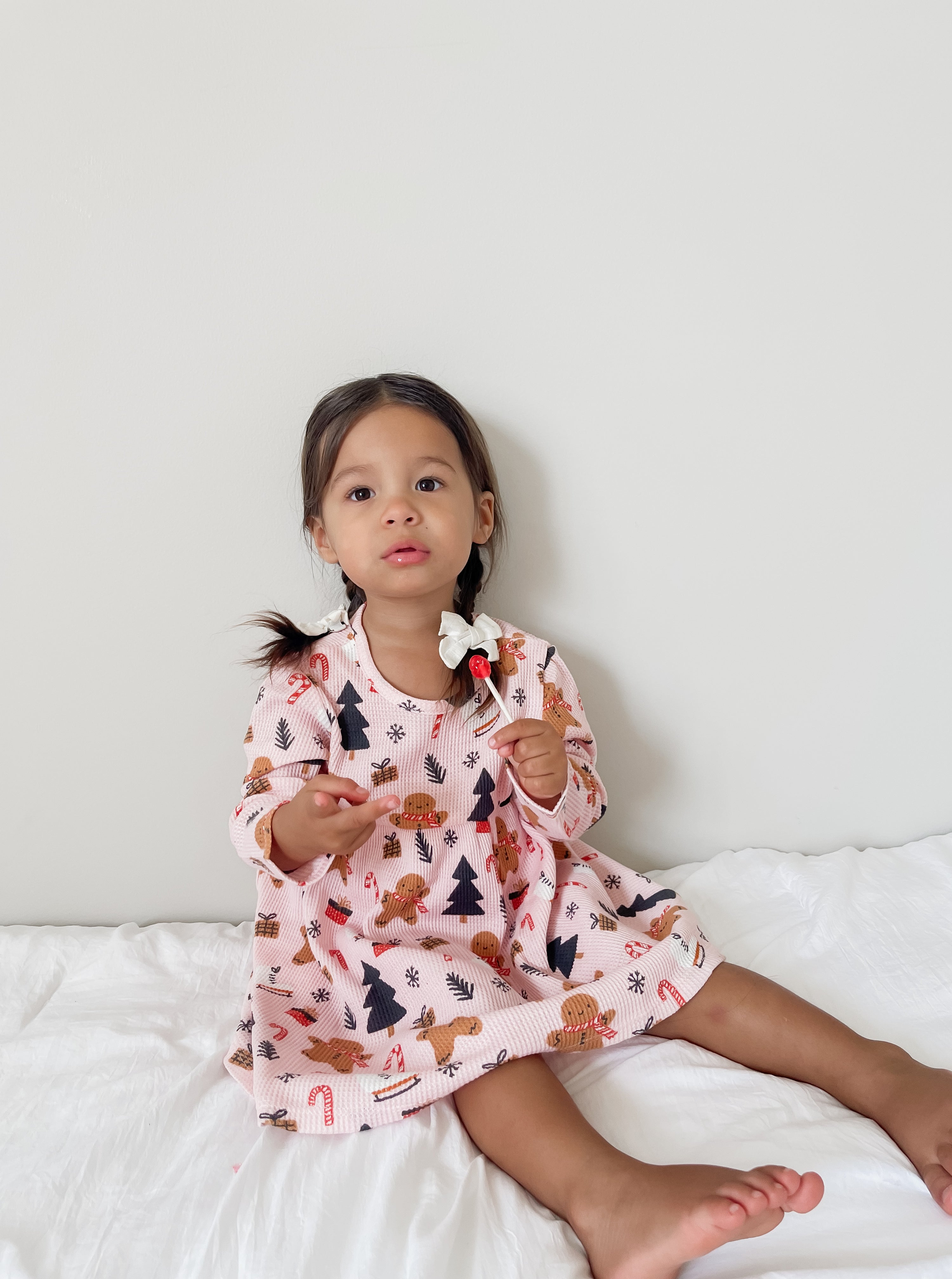 Young child in festive dress, sitting on a white blanket, holding a lollipop, with a neutral background.
